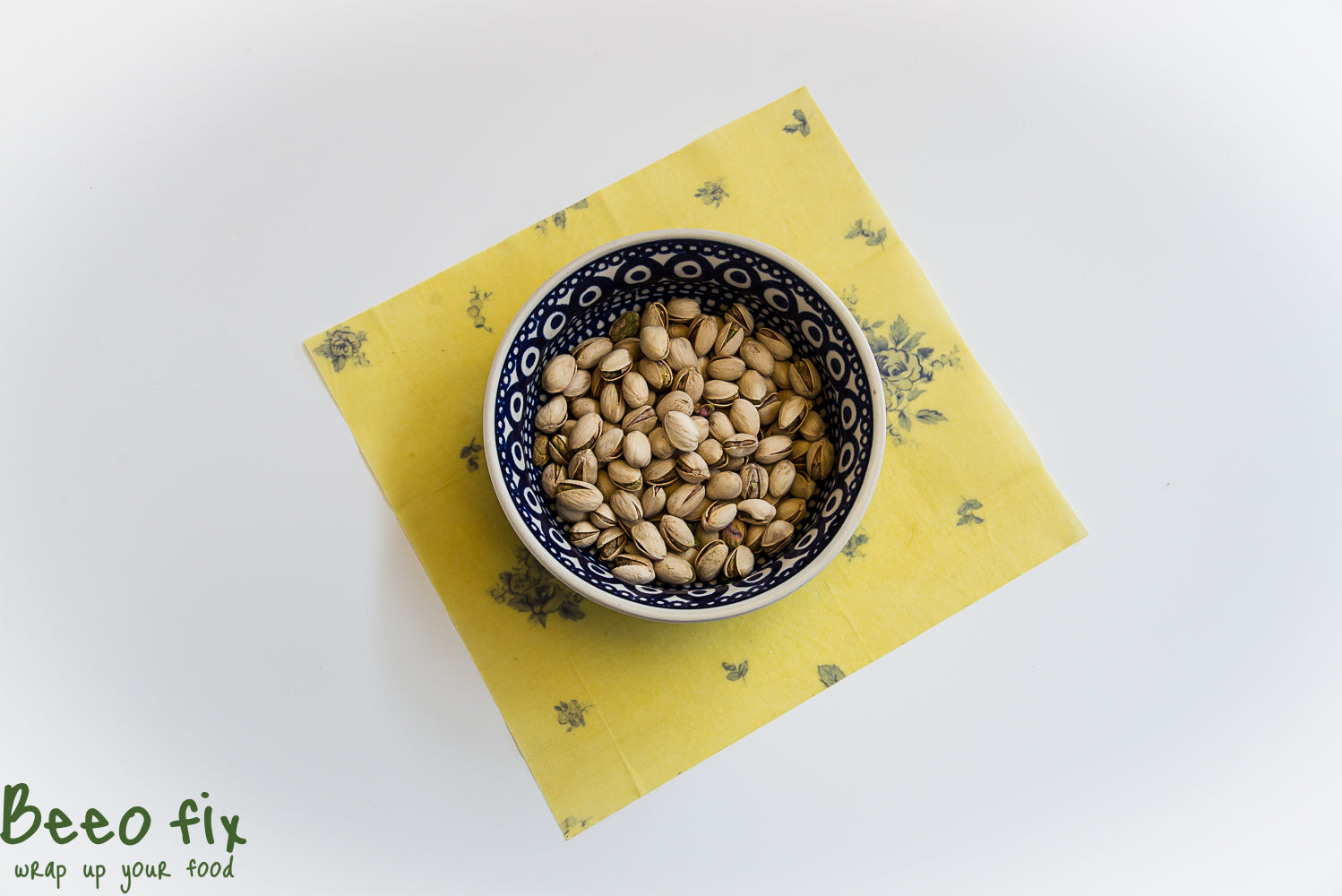 Blaue Schüssel mit Pistazien auf gelb gemustertem Tuch, darunter der Text "Beeo fix - wrap up your food".