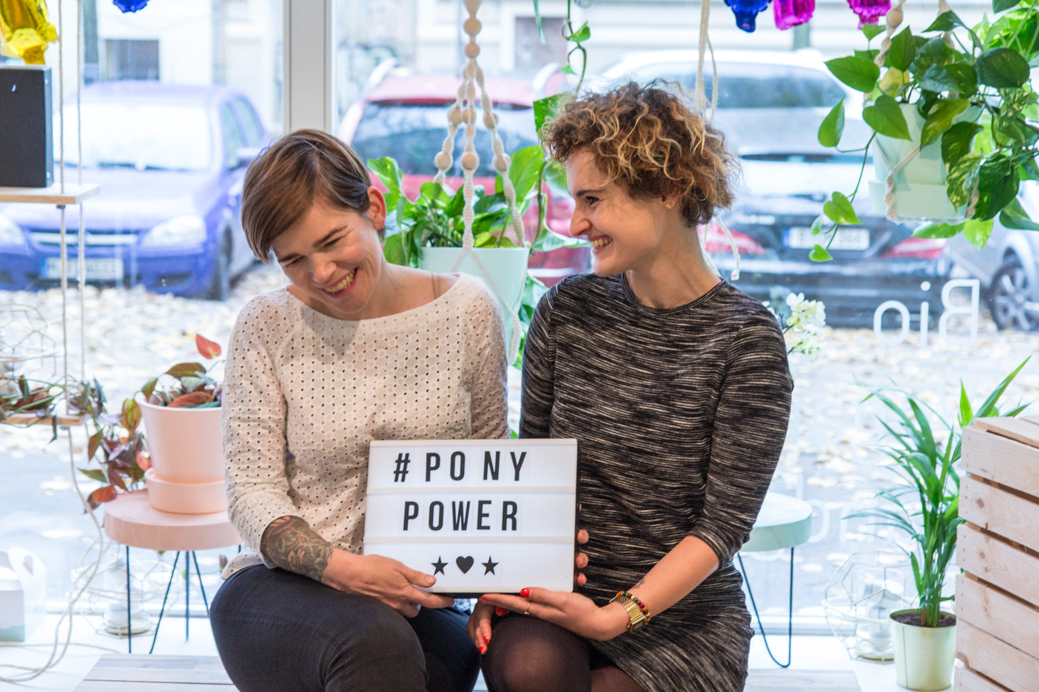 Zwei lachende Personen sitzen nebeneinander und halten ein Schild mit der Aufschrift "#PONY POWER" umgeben von Pflanzen.