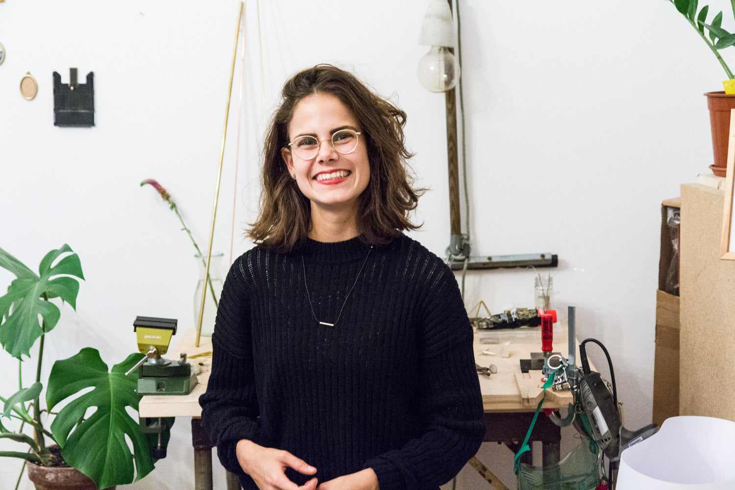 Lächelnde Person mit Brille in einem Atelier mit Pflanzen und Werkzeugen im Hintergrund.