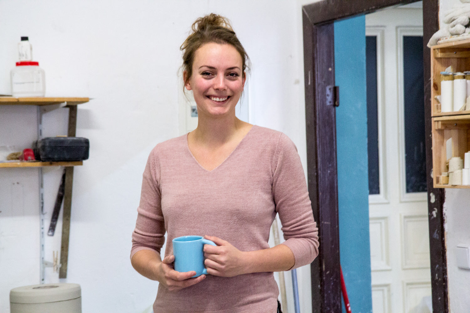 Eine lächelnde Person hält eine blaue Tasse in einem Raum mit Regalen und einer Tür im Hintergrund.