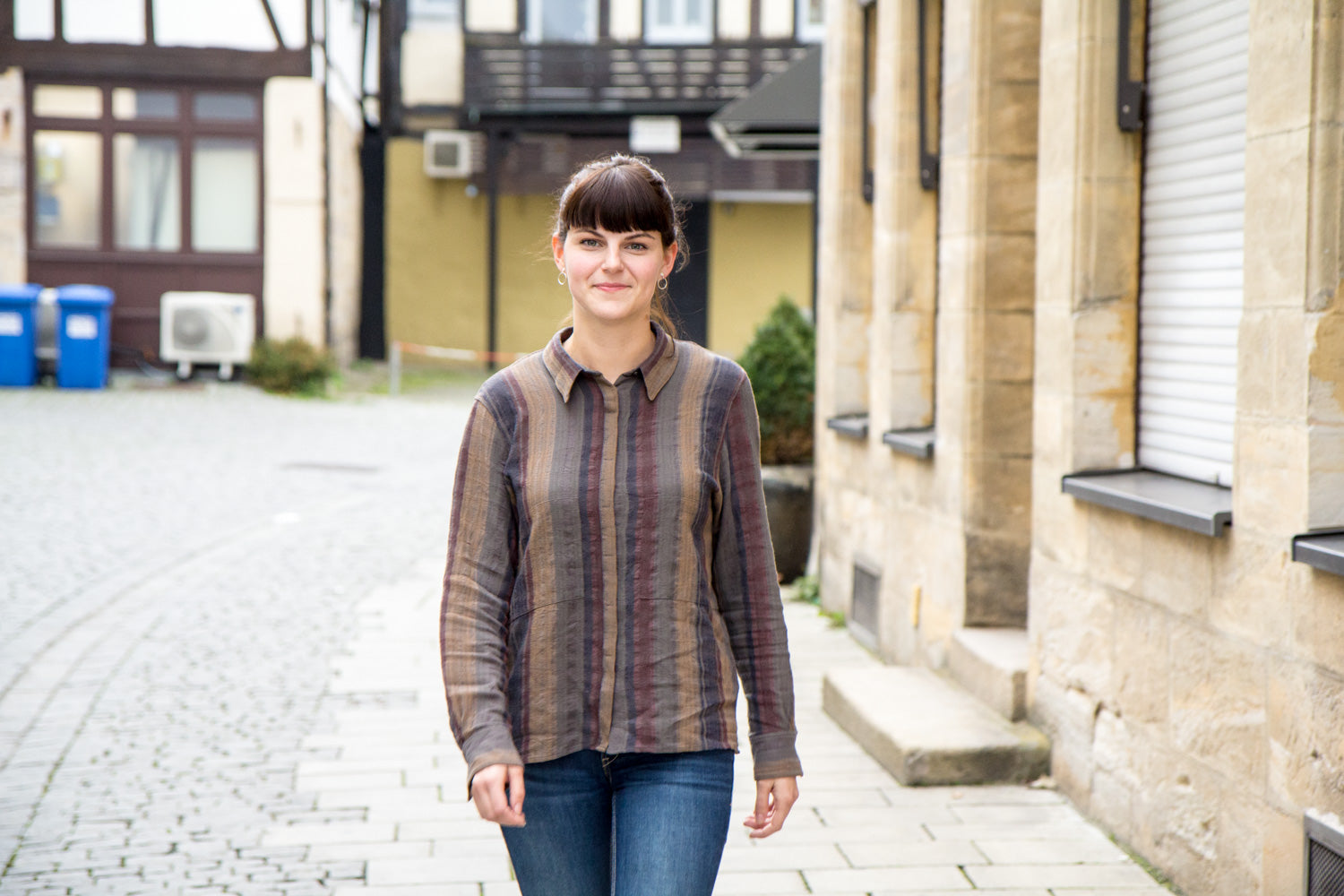 Eine Frau geht lächelnd auf einem gepflasterten Gehweg entlang einer Gebäudefassade spazieren.