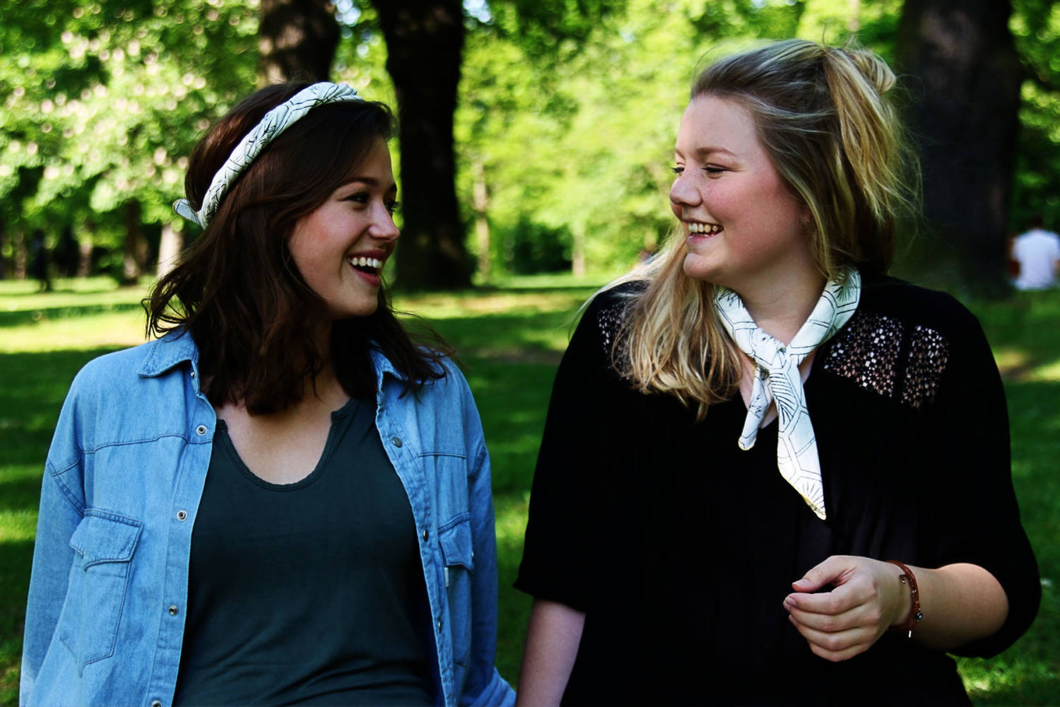 Zwei lachende Frauen stehen nah beieinander im Park, umgeben von Bäumen und einer grasbewachsenen Fläche.