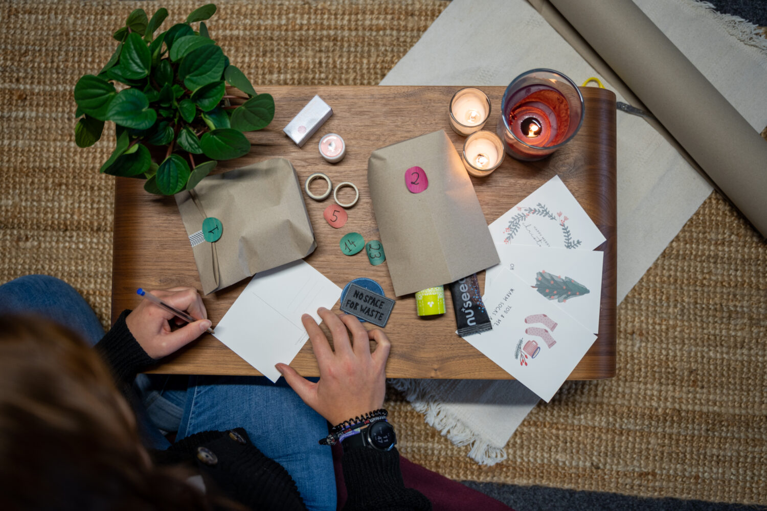 Person schreibt Karte auf Tisch mit Geschenken, Kerzen, Karten, Pflanzen und Verpackungspapier.