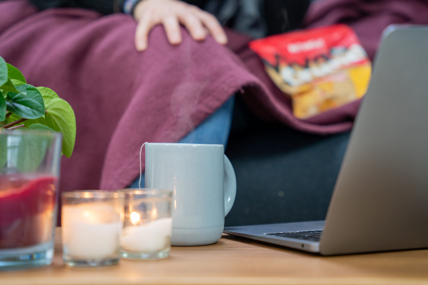 Eine Szene mit Laptop, Tasse Tee, Kerzen und Pflanze auf einem Tisch, daneben eine Person mit Decke und Snackpackung.