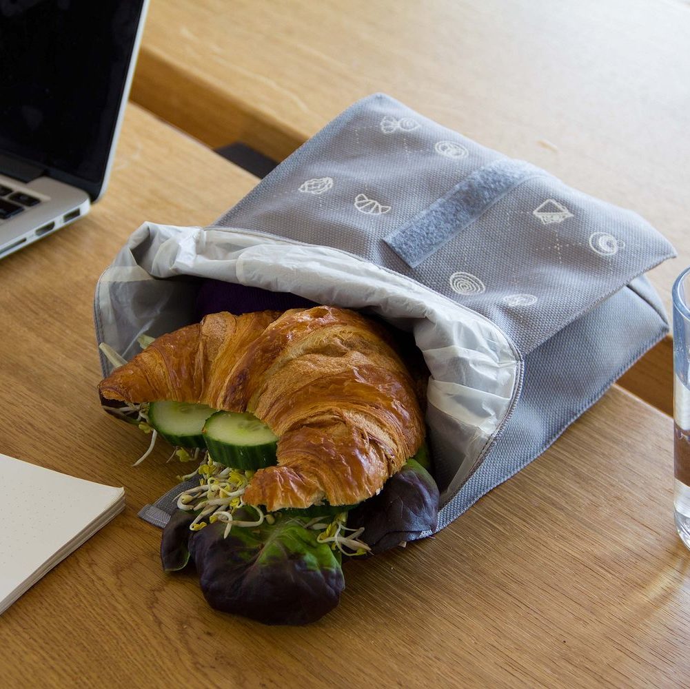 Ein Croissant-Sandwich mit Salat, Gurken und Sprossen in einem grauen Beutel liegt auf einem Holztisch neben einem Laptop.