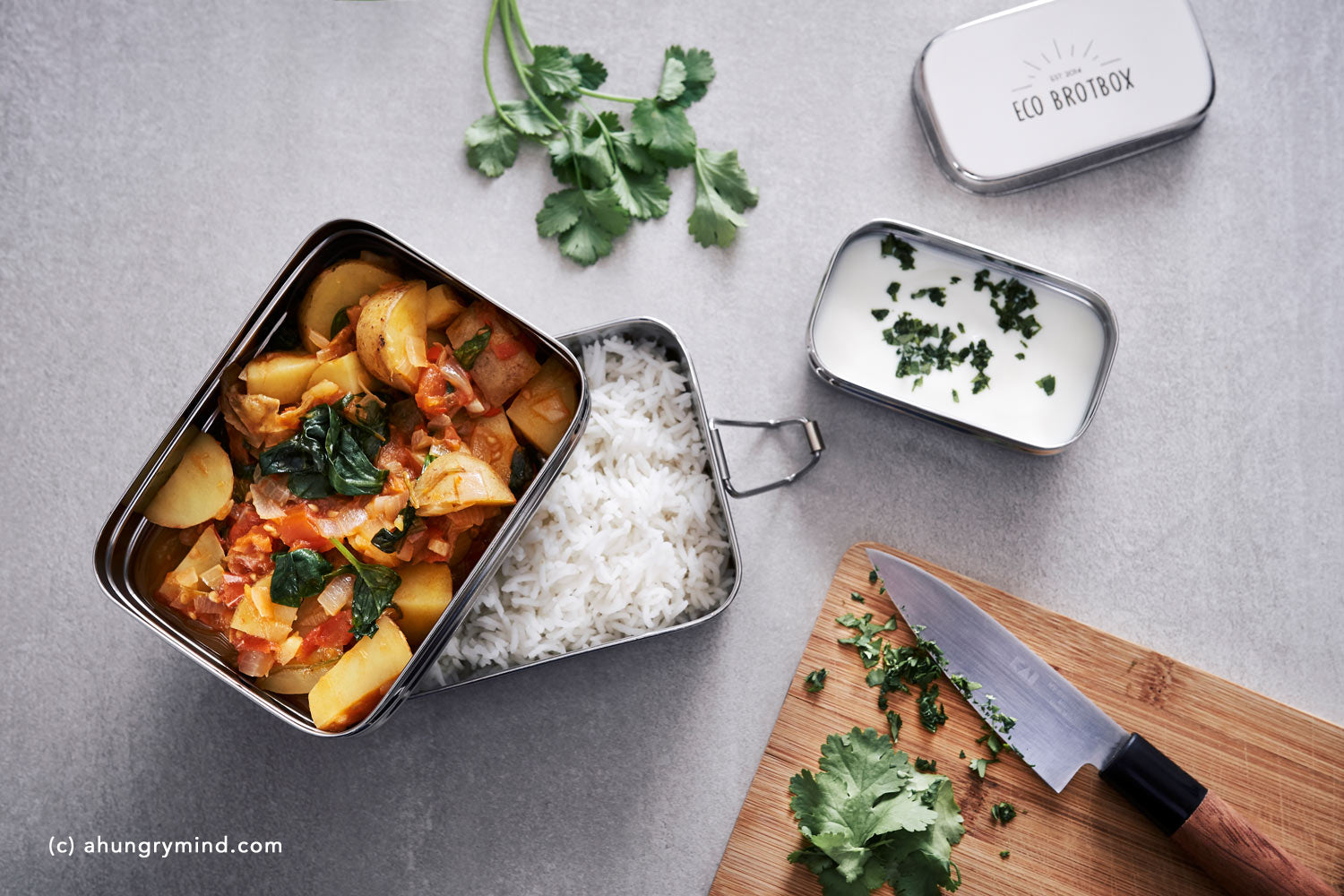 Ein Lunchbox-Menü mit Kartoffelcurry und Reis, daneben Joghurt mit Kräutern und ein Schneidebrett mit Messer und Koriander.