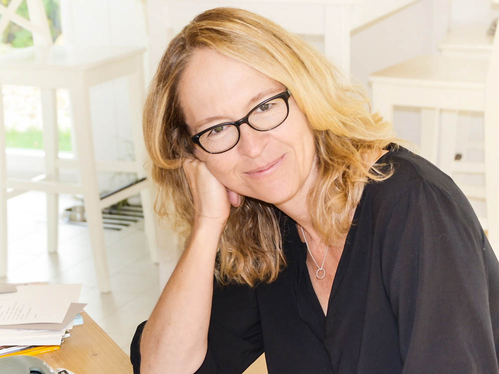 Eine lächelnde Frau mit Brille sitzt an einem Tisch und stützt den Kopf auf ihre Hand.