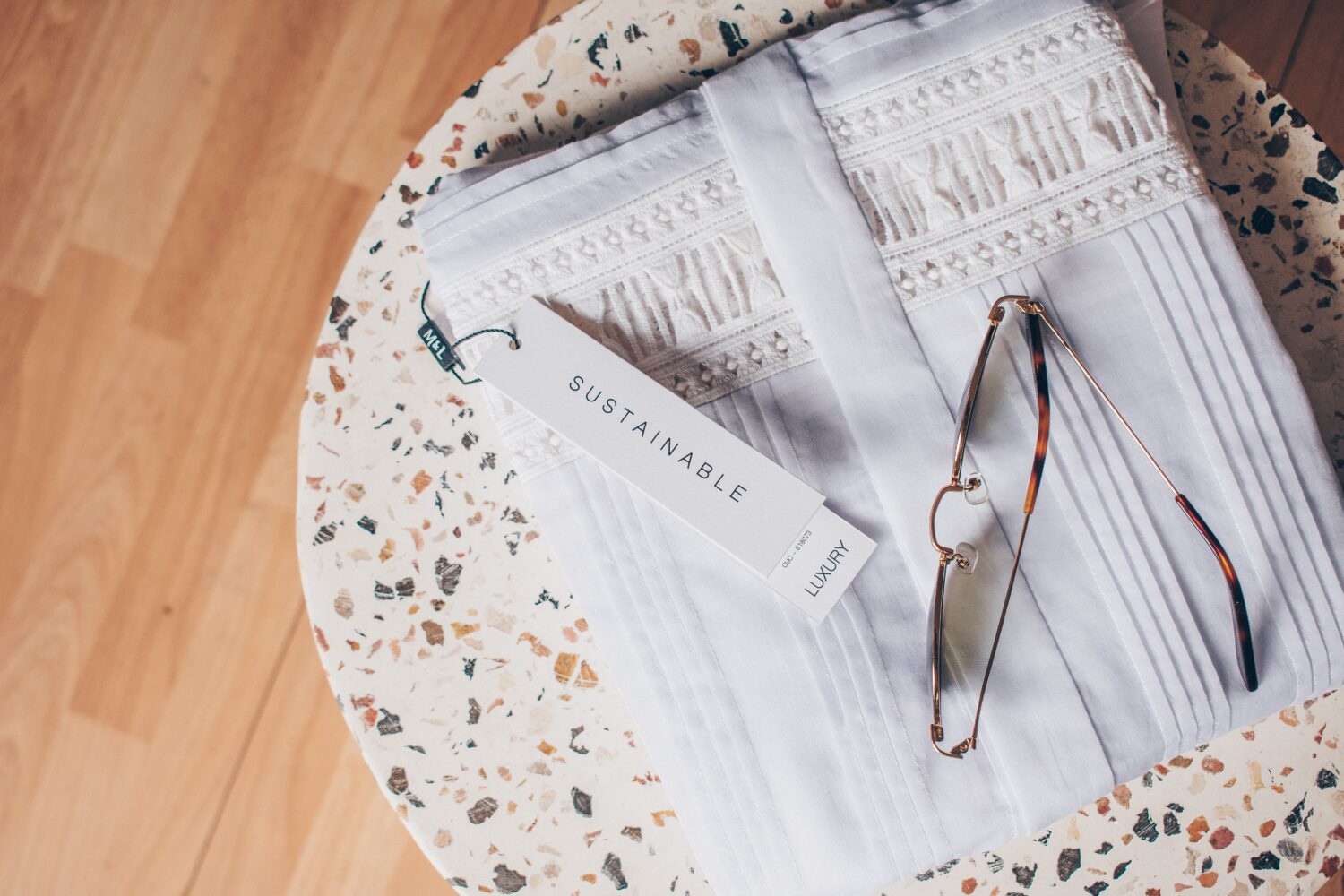 Weiße gefaltete Kleidung mit einem Schild "Sustainable Luxury" und einer Brille auf einem Tisch mit Terrazzo-Oberfläche.