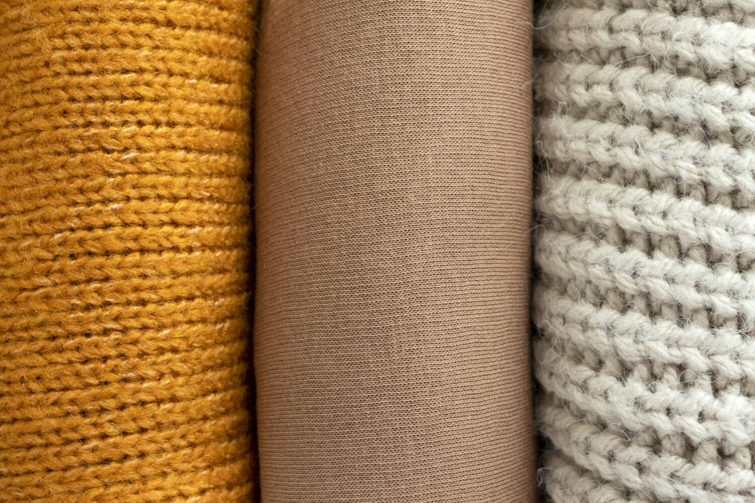 Drei Rollen Stoff nebeneinander: gelber Strick, beige Webstoff und grauer Strick.