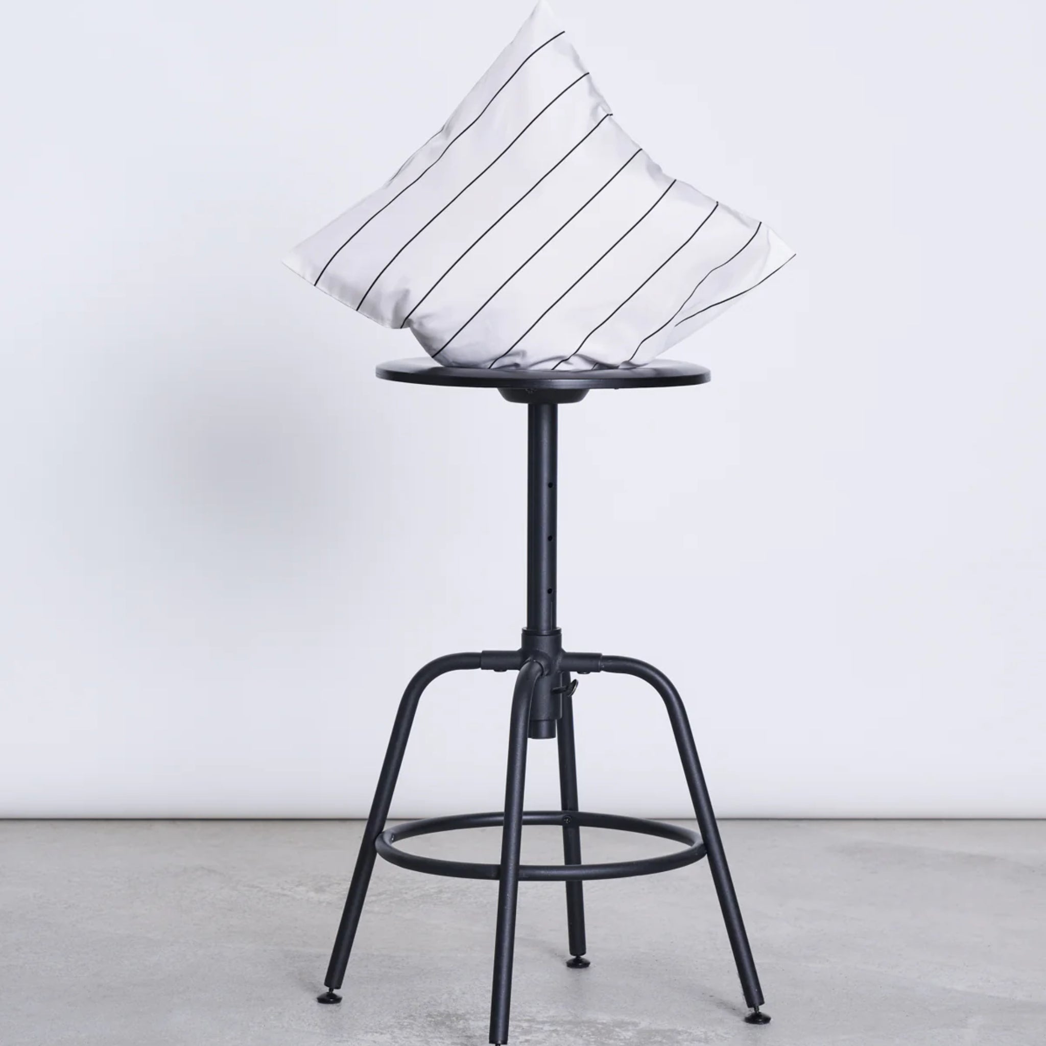 Ein schwarzer Metallhocker mit einem weiß-gestreiften Kissen auf der Sitzfläche steht vor einer grauen Wand.