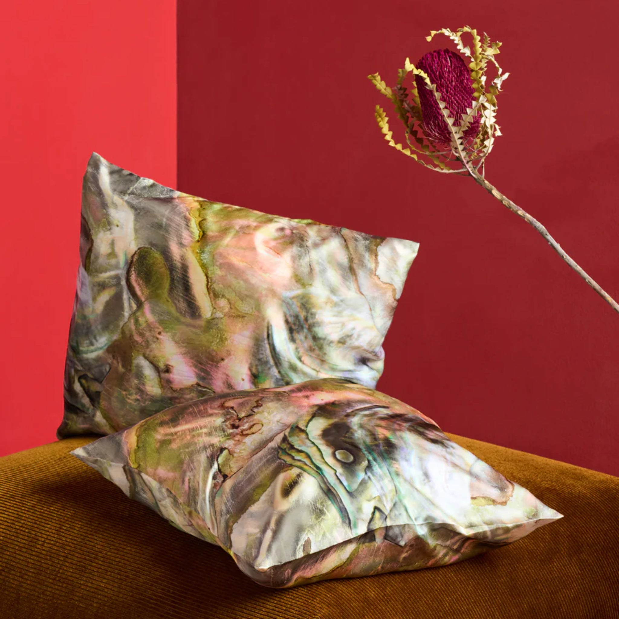 Zwei bunte Kissen mit marmoriertem Muster liegen auf einem braunen Sofa vor einer roten Wand; im Hintergrund eine Blume.