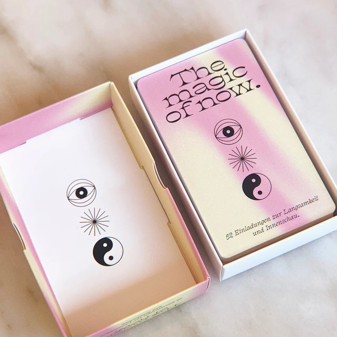 Eine Schachtel mit einem Kartenspiel mit der Aufschrift The magic of now. in rosa und beigen Farbverläufen, mit Yin-Yang und Augensymbolen auf den Karten. Die Schachtel enthält einen deutschen Text: 52 Einladungen zur Langsamkeit und Innenschau.