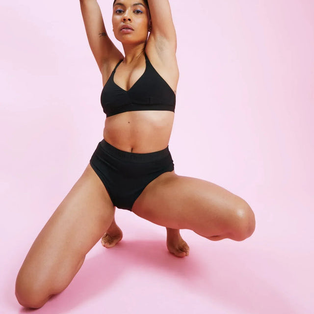 Eine Frau, die einen schwarzen Sport-BH und eine hoch taillierte Hose trägt, posiert selbstbewusst vor einem rosafarbenen Hintergrund, kniet auf einem Knie und hebt die Arme über den Kopf.