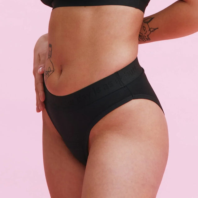 Eine Person, die einen schwarzen BH und schwarze Unterwäsche trägt, steht vor einem rosafarbenen Hintergrund und hat eine Hand auf die Hüfte gestützt. Auf ihrem Arm und Oberkörper sind mehrere Tätowierungen zu sehen.