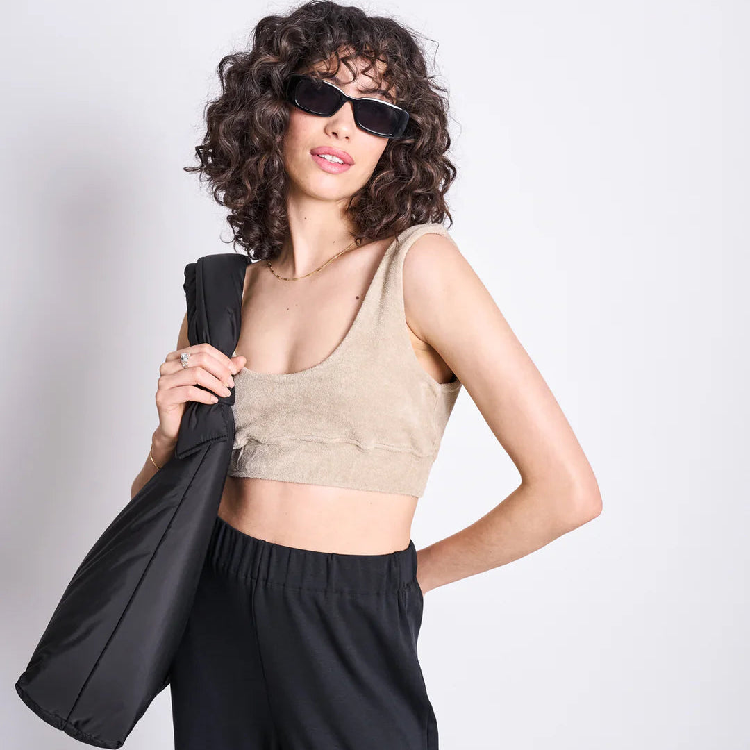 Frau mit lockigem Haar trägt Sonnenbrille, beiges Oberteil und hält schwarze Jacke über Schulter, schaut zur Kamera.