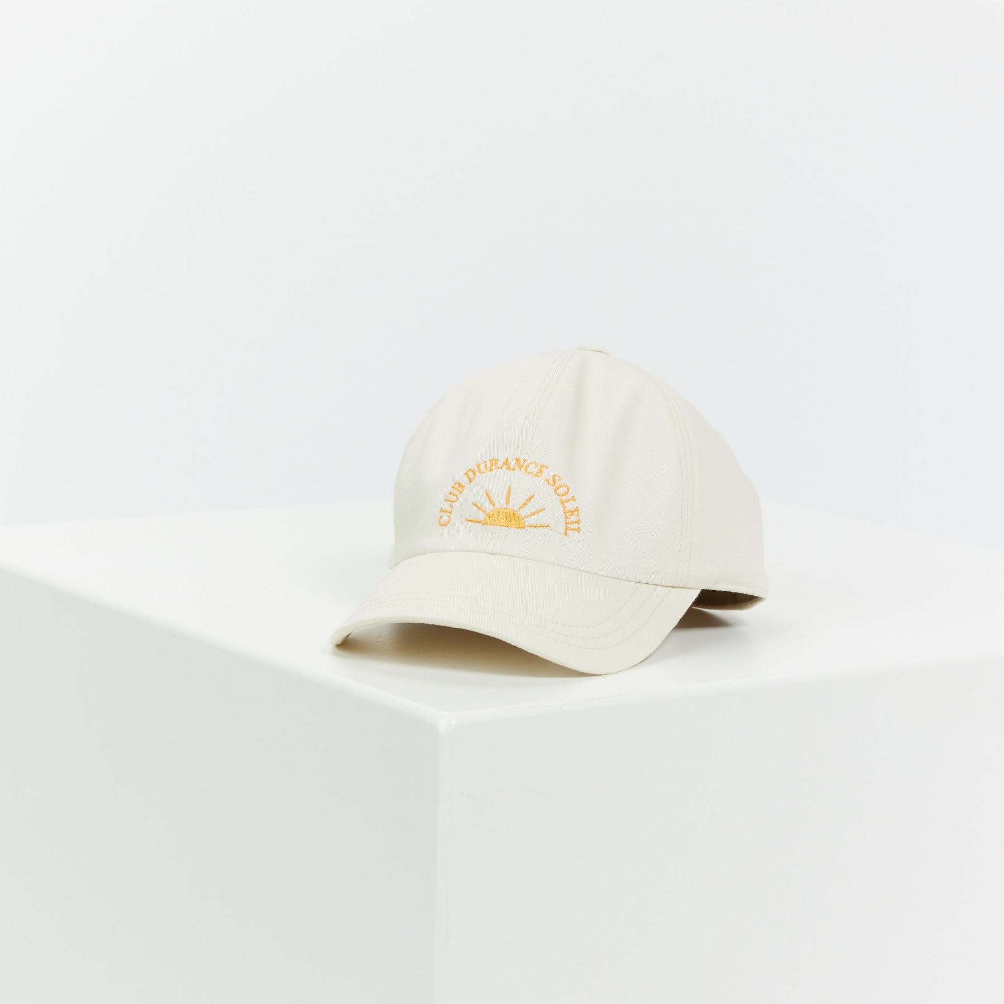 Beige Basecap mit der Aufschrift "Club Durancé Soleil" und einem gestickten Sonnen-Design liegt auf einem weißen Podest.