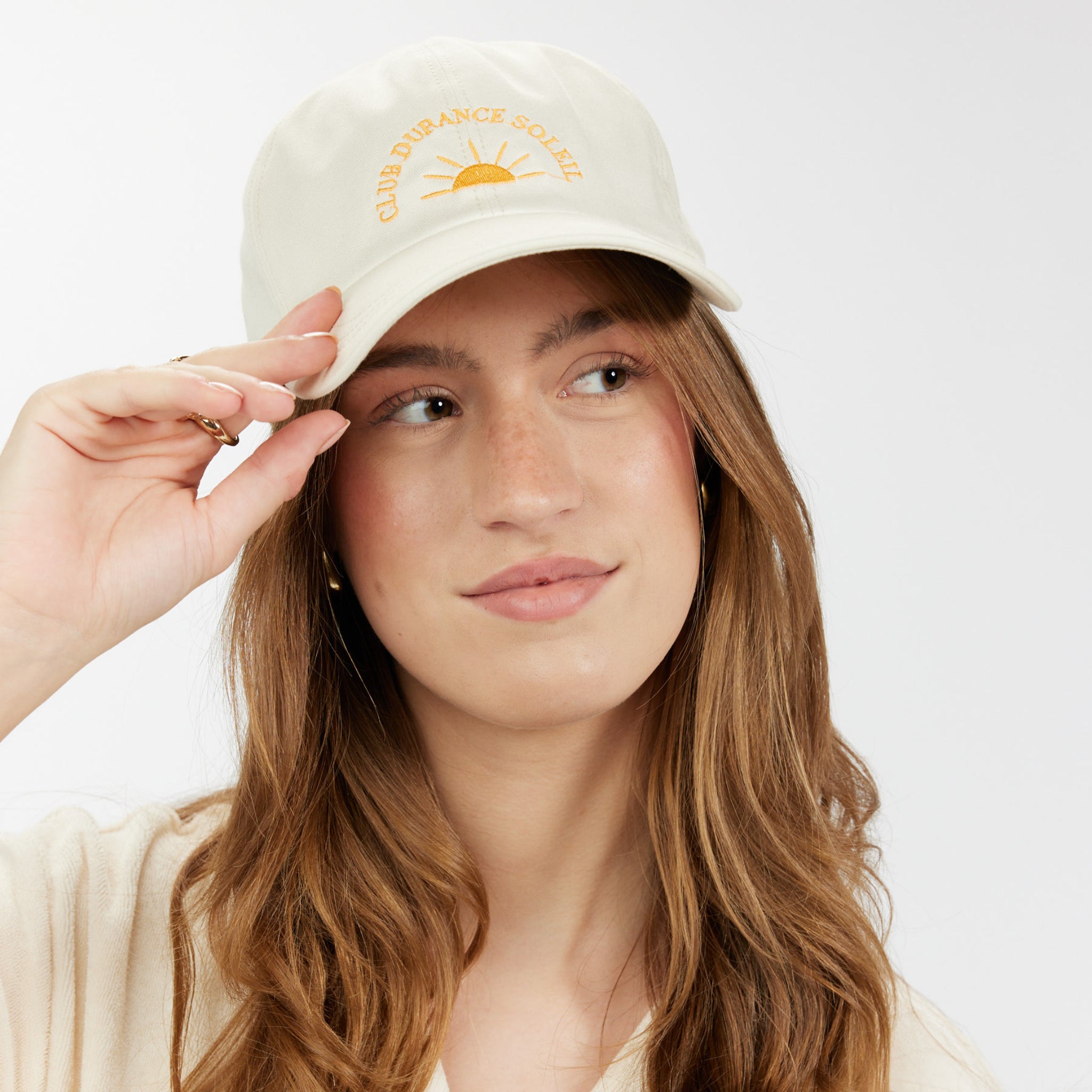 Frau mit langerem Haar trägt eine beigefarbene Kappe mit Sonnendesign und lächelndem Gesichtsausdruck.