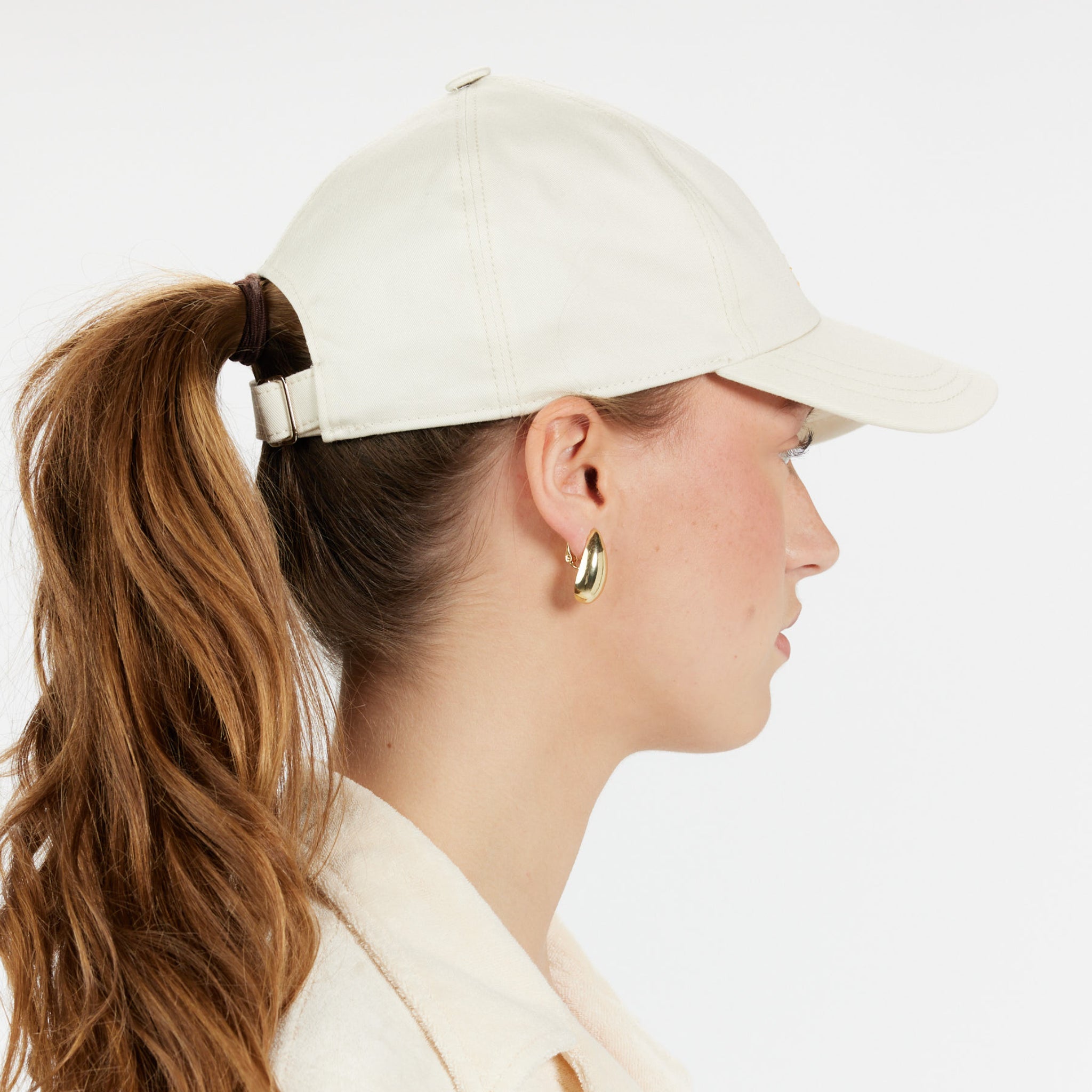 Frau mit weißer Basecap und langen braunen Haaren im Pferdeschwanz, trägt goldene Ohrringe, Seitenprofilansicht.