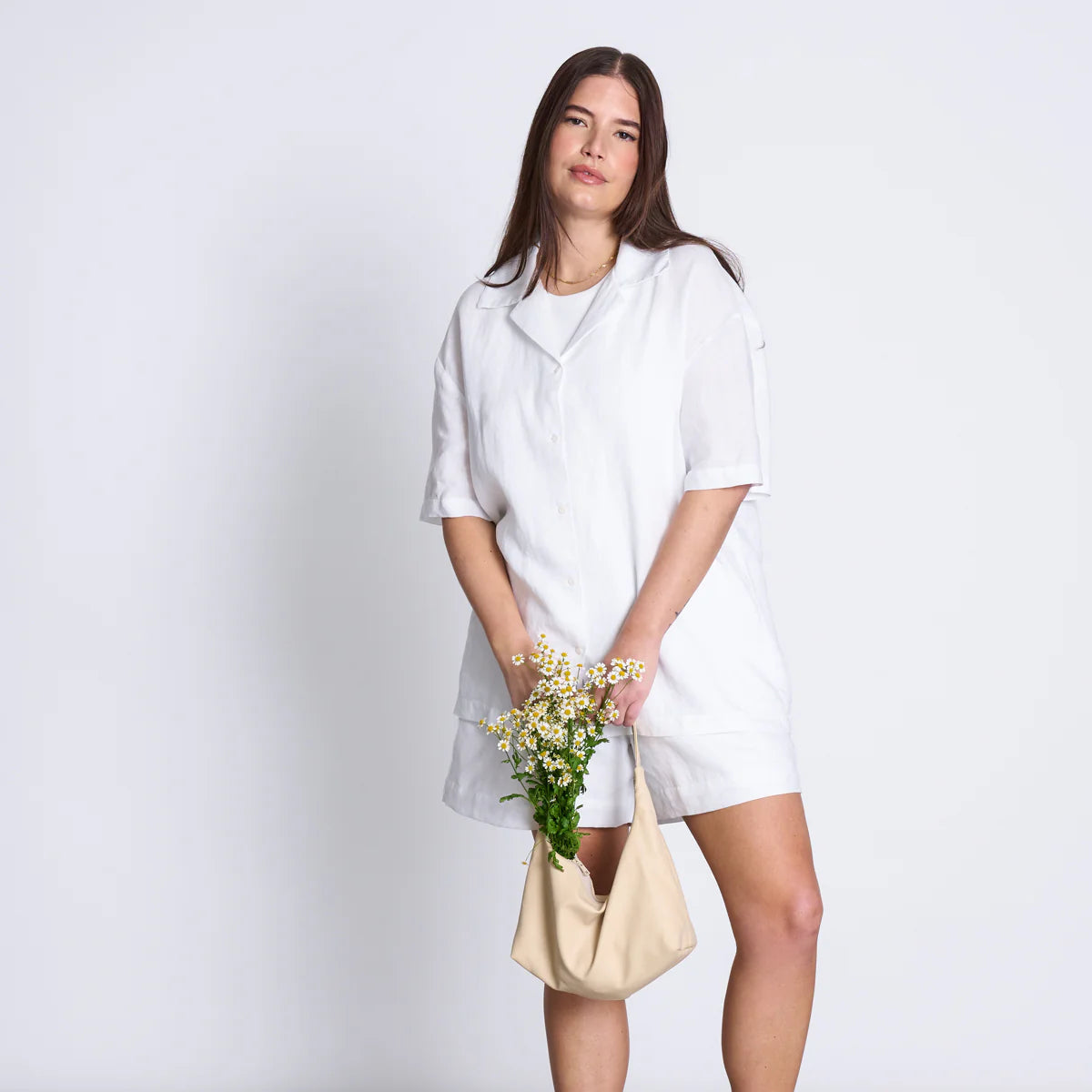 Frau in weißem Kleid hält eine Tasche mit kleinen weißen Blumen vor einem weißen Hintergrund.