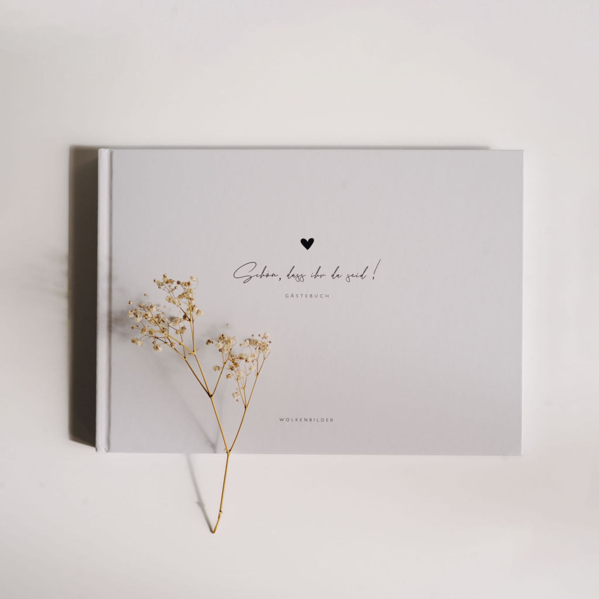 Gästebuch mit der Aufschrift „Schön, dass du da bist!“ und einem kleinen Herz, dekoriert mit einem Zweig Trockenblumen.
