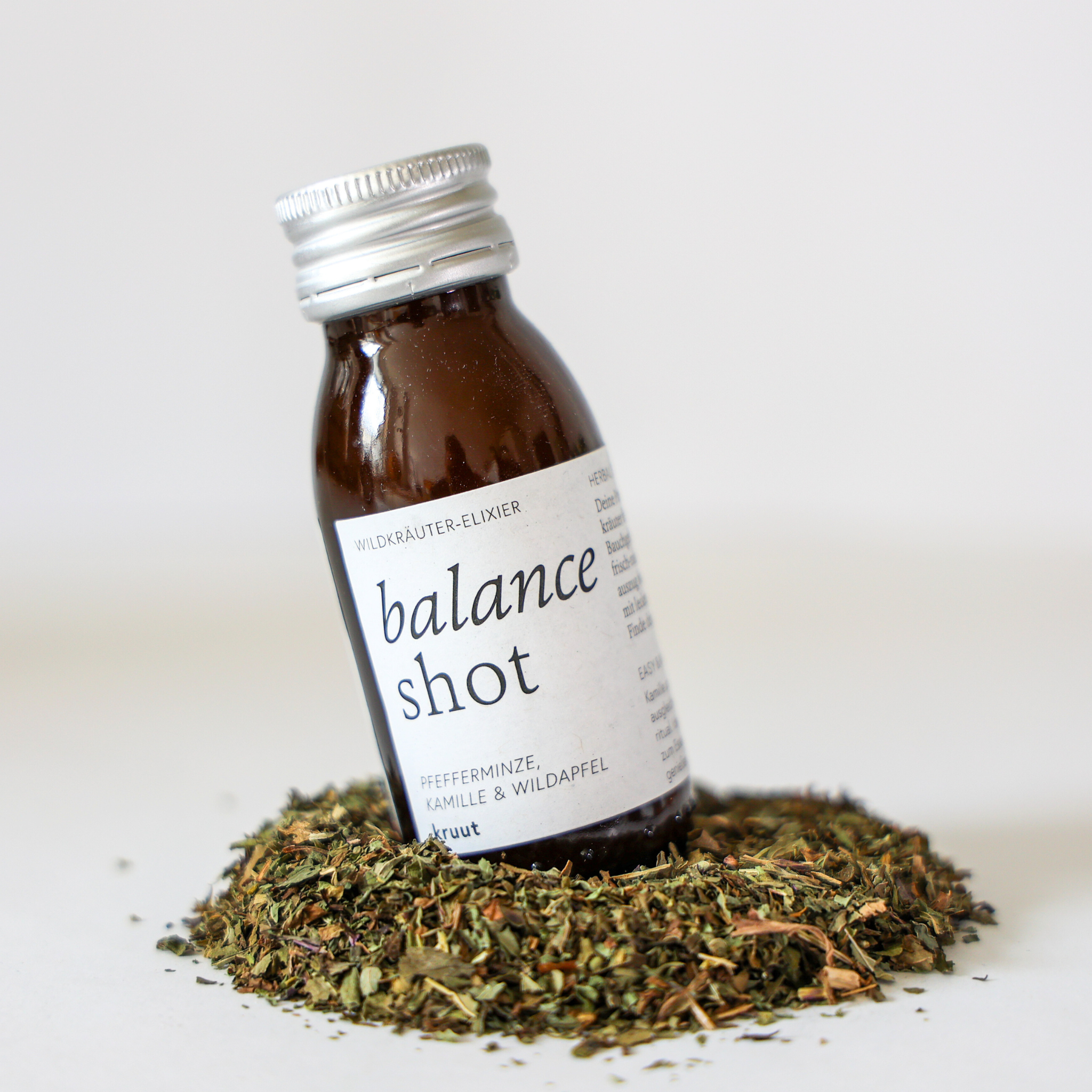 Braunes Fläschchen mit der Aufschrift "Balance Shot" in einem Kreis aus getrockneten Kräutern auf weißem Hintergrund.