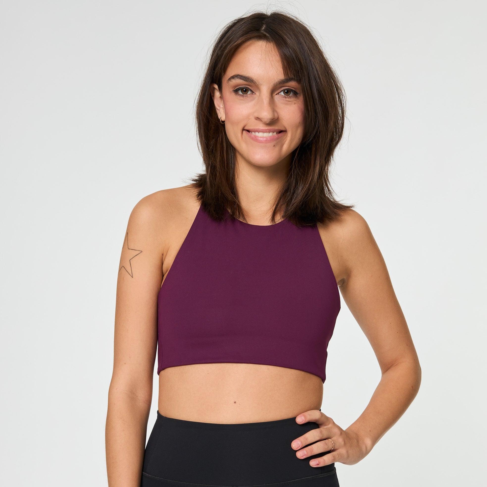 Frau in lila Sporttop lächelt, hat eine Stern-Tätowierung auf dem Arm, vor weißem Hintergrund.