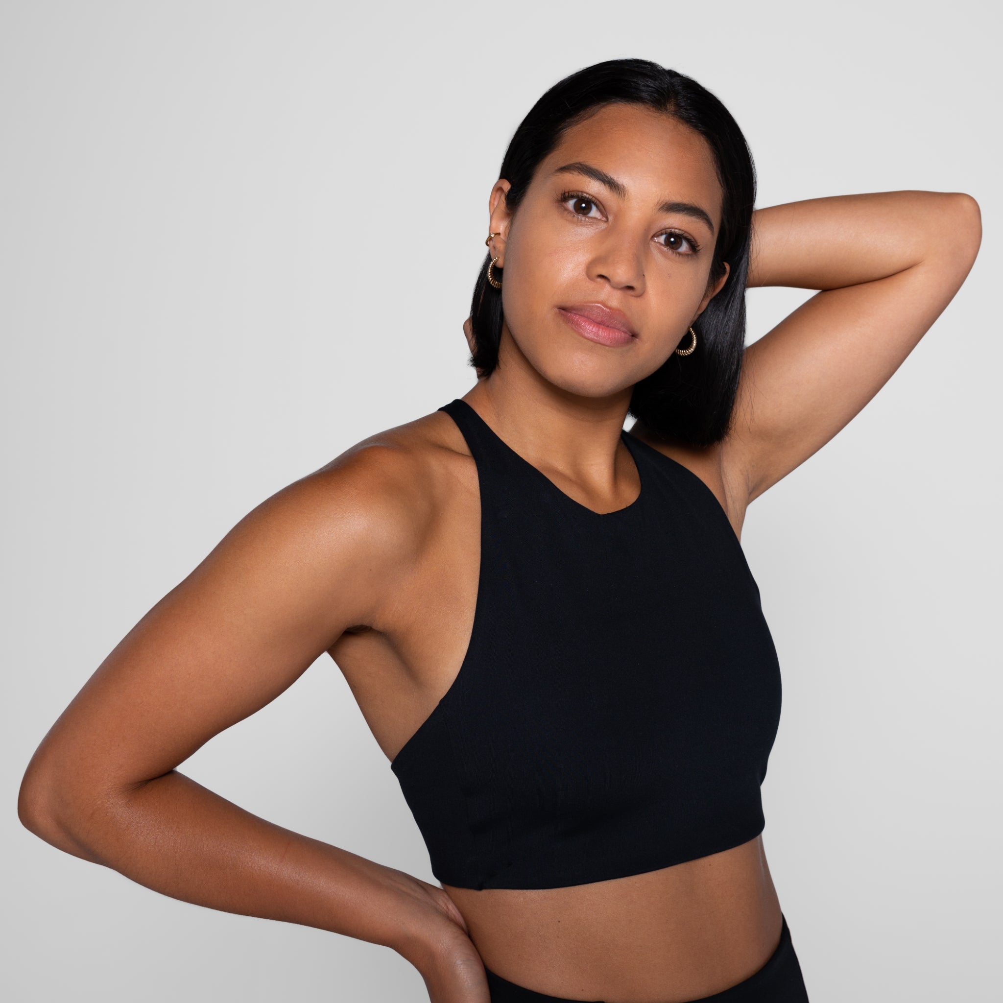 Person im schwarzen Tanktop posiert vor grauem Hintergrund mit einer Hand am Kopf und einem leichten Lächeln.