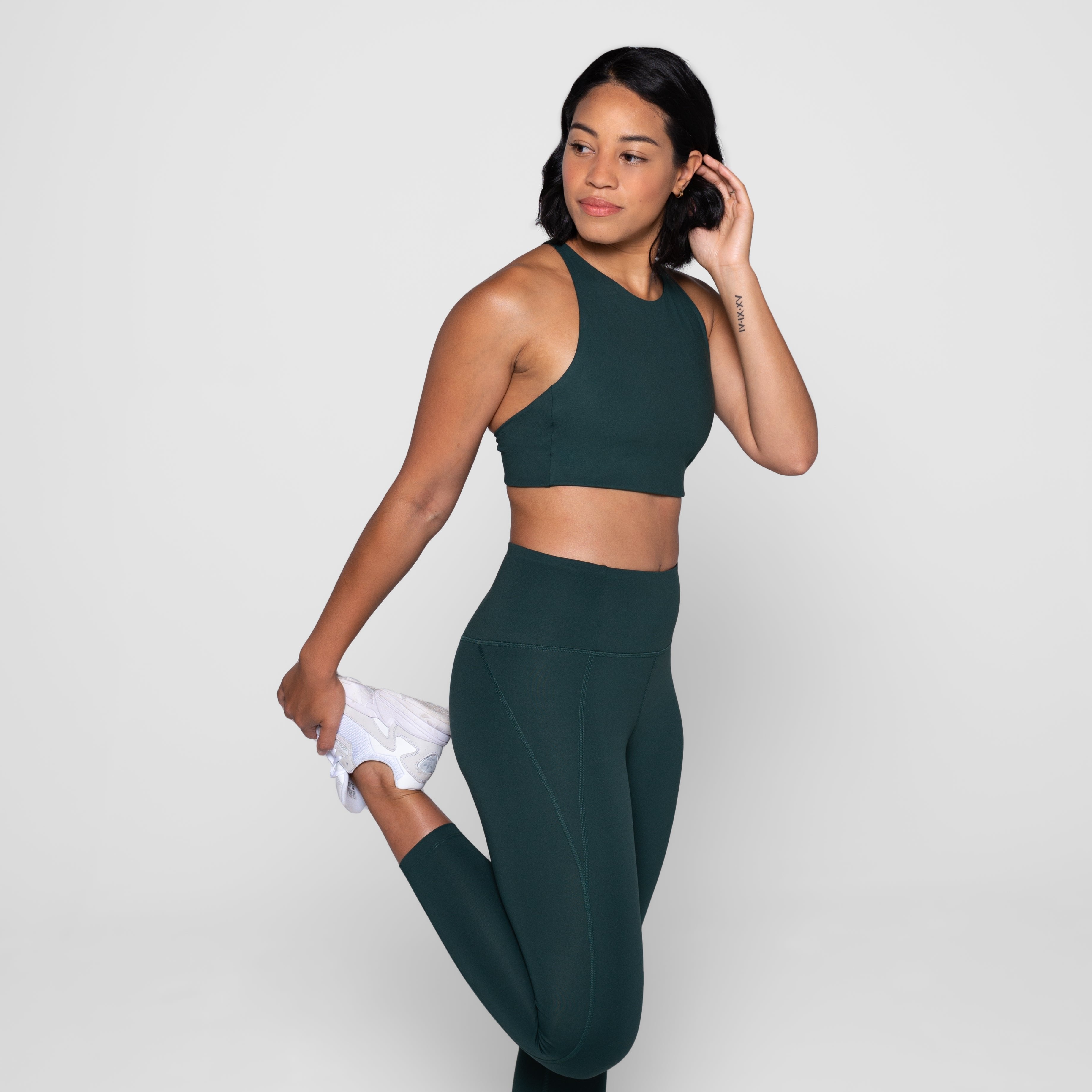 Frau in dunkelgrüner Sportkleidung dehnt ihr Bein, während sie lächelt und zur Seite schaut, vor grauem Hintergrund.