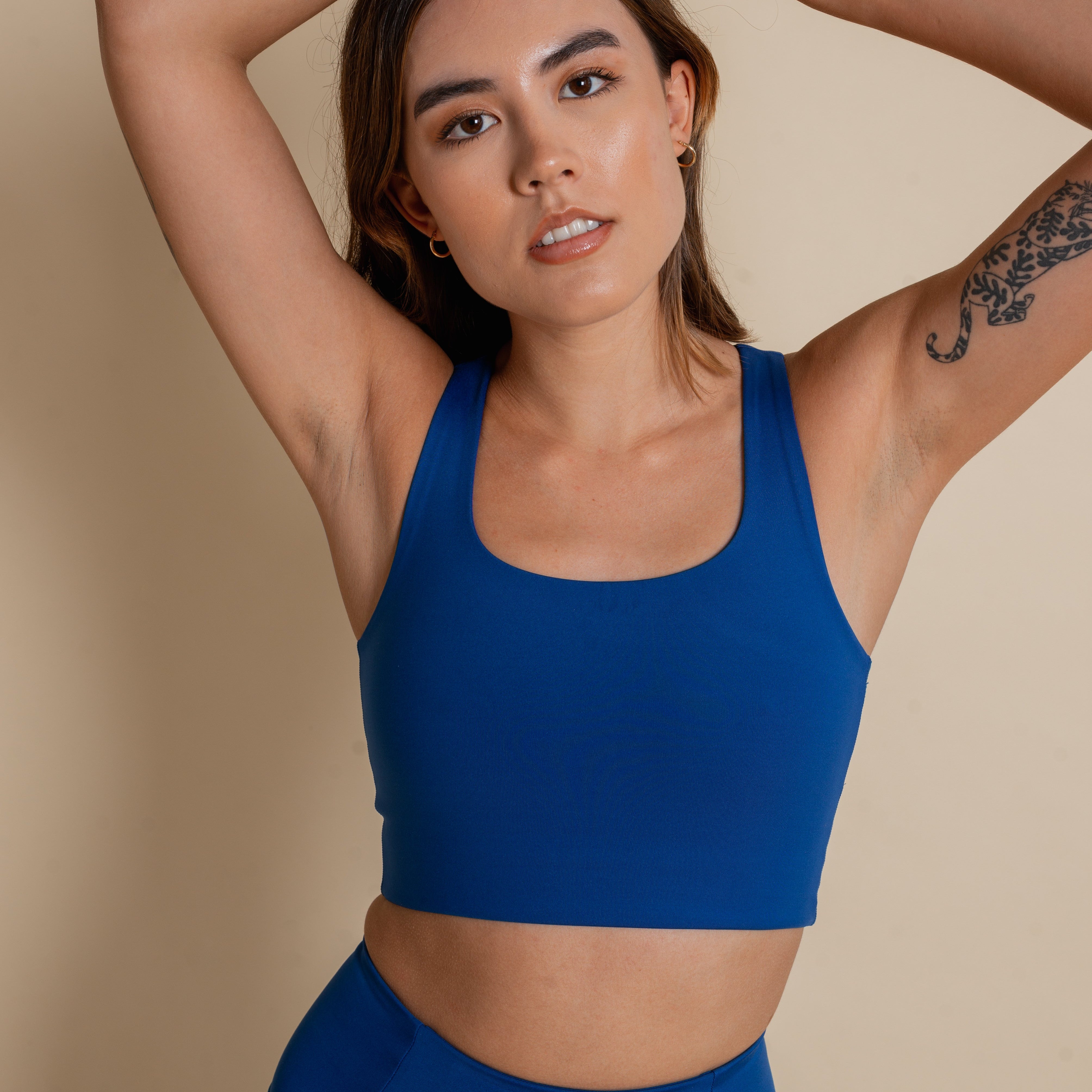 Eine Frau mit langen braunen Haaren, die einen blauen Sport-BH und passende Leggings trägt, steht mit über den Kopf erhobenen Armen vor einem beigen Hintergrund. Sie hat eine schwarze Tätowierung auf ihrem linken Oberarm.