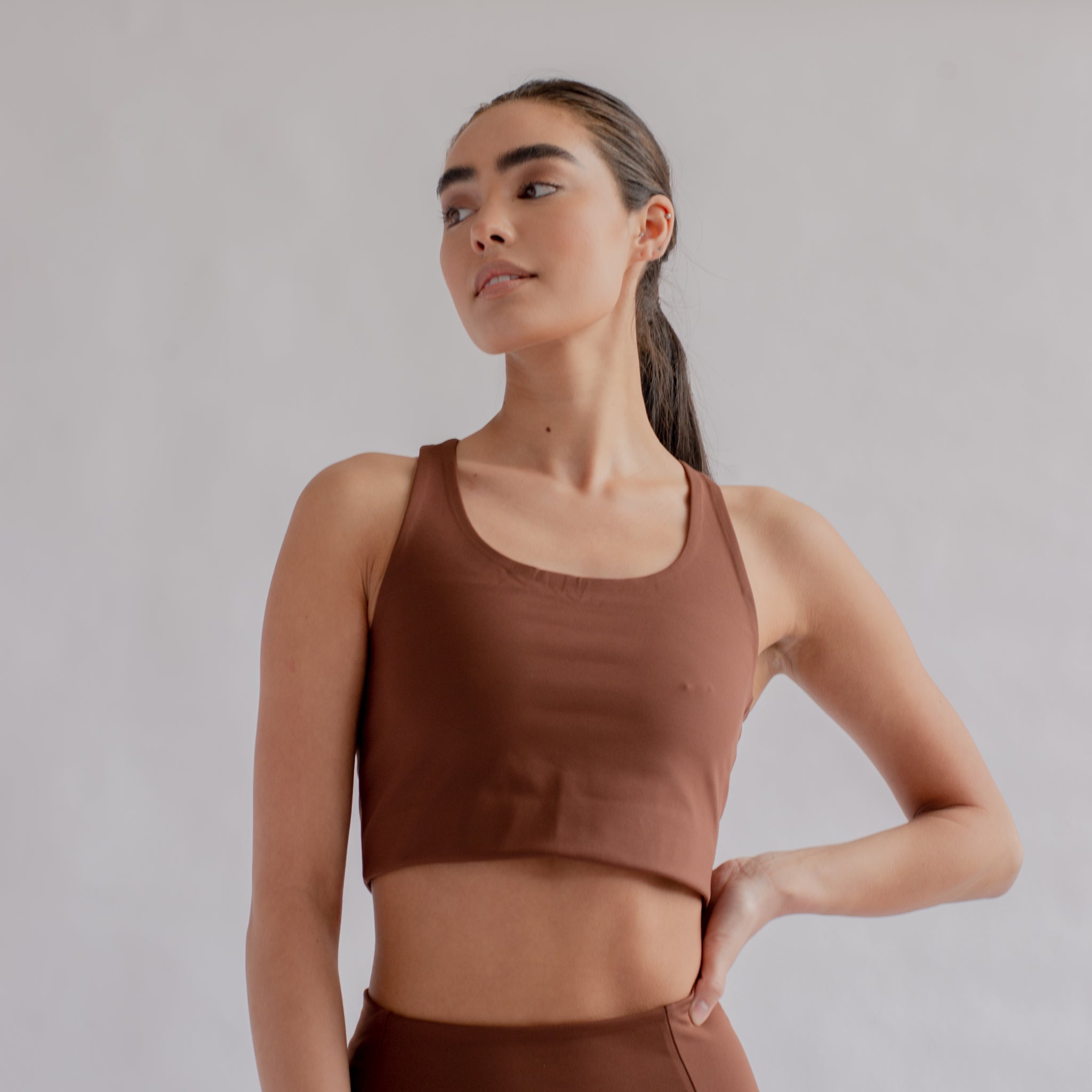 Junge Frau in braunem Sporttop und Leggings, steht mit einer Hand in der Hüfte in einem minimalistischen Raum.