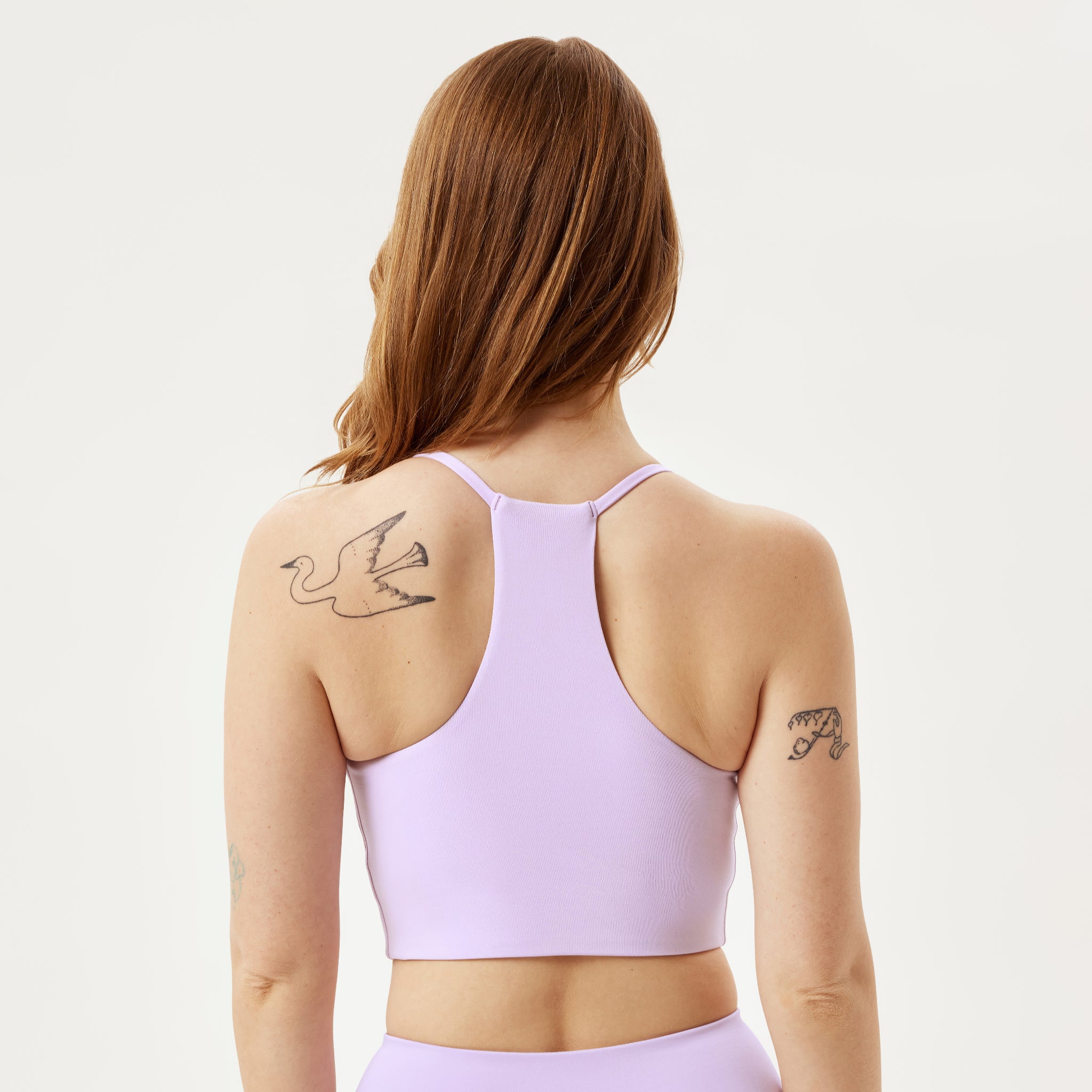 Man sieht eine Frau mit langen braunen Haaren von hinten, die einen hellvioletten Sport-BH trägt. Sie hat ein Vogeltattoo auf der linken Schulter und ein kleines Texttattoo auf dem rechten Arm. Der Hintergrund ist einfarbig und hell.
