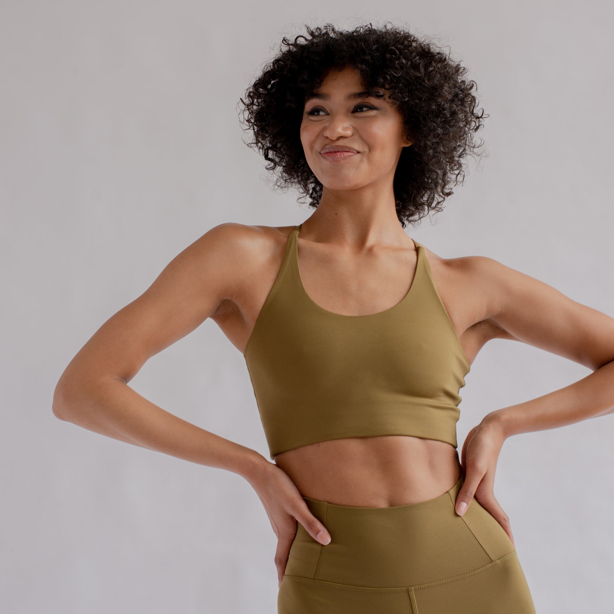 Frau in olivgrünem Sport-BH und Leggings lächelt mit den Händen in der Hüfte vor neutralem Hintergrund.