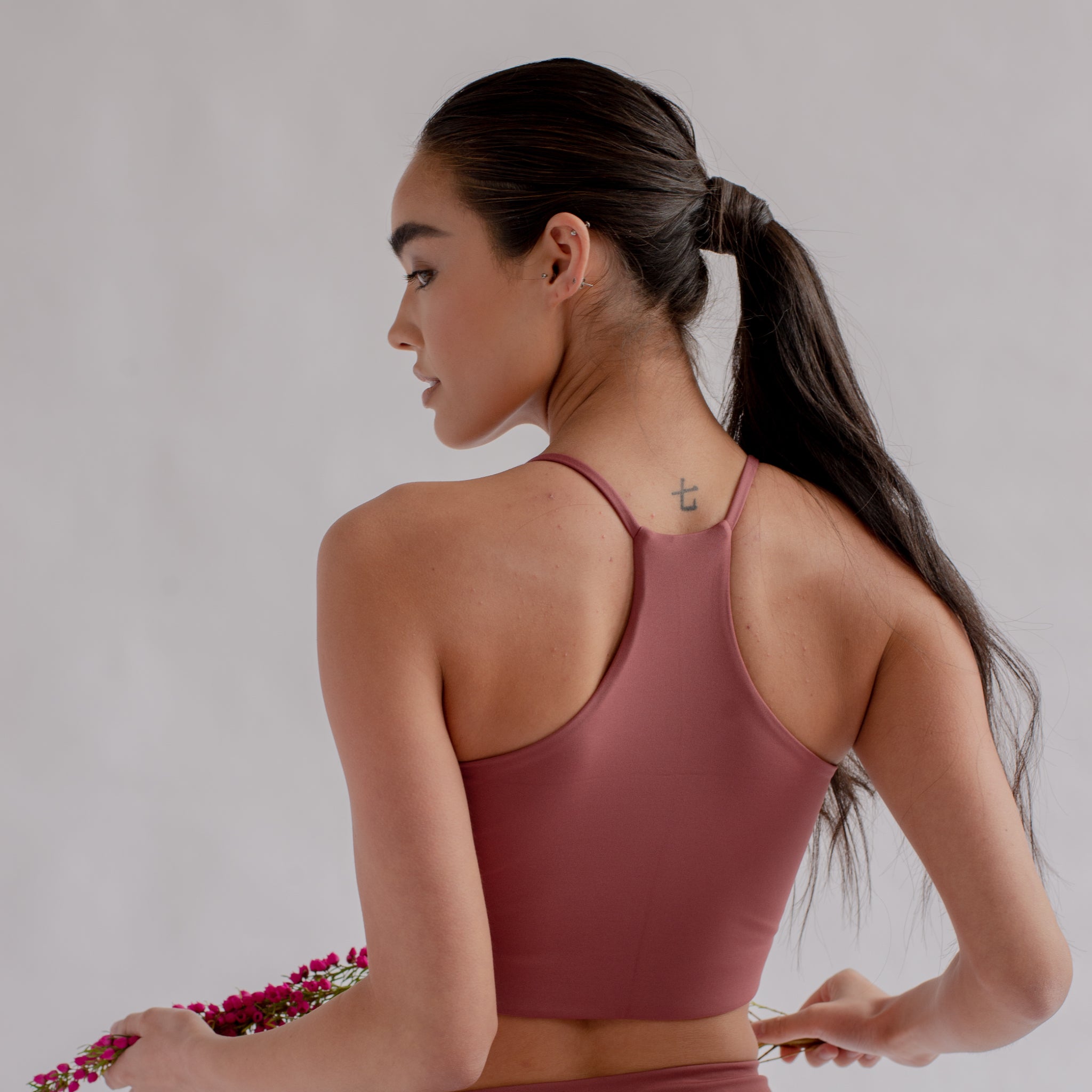 Frau in rosa Sporttop zeigt Tätowierung im Nacken und hält rosa Blumen, seitlich beleuchtet.