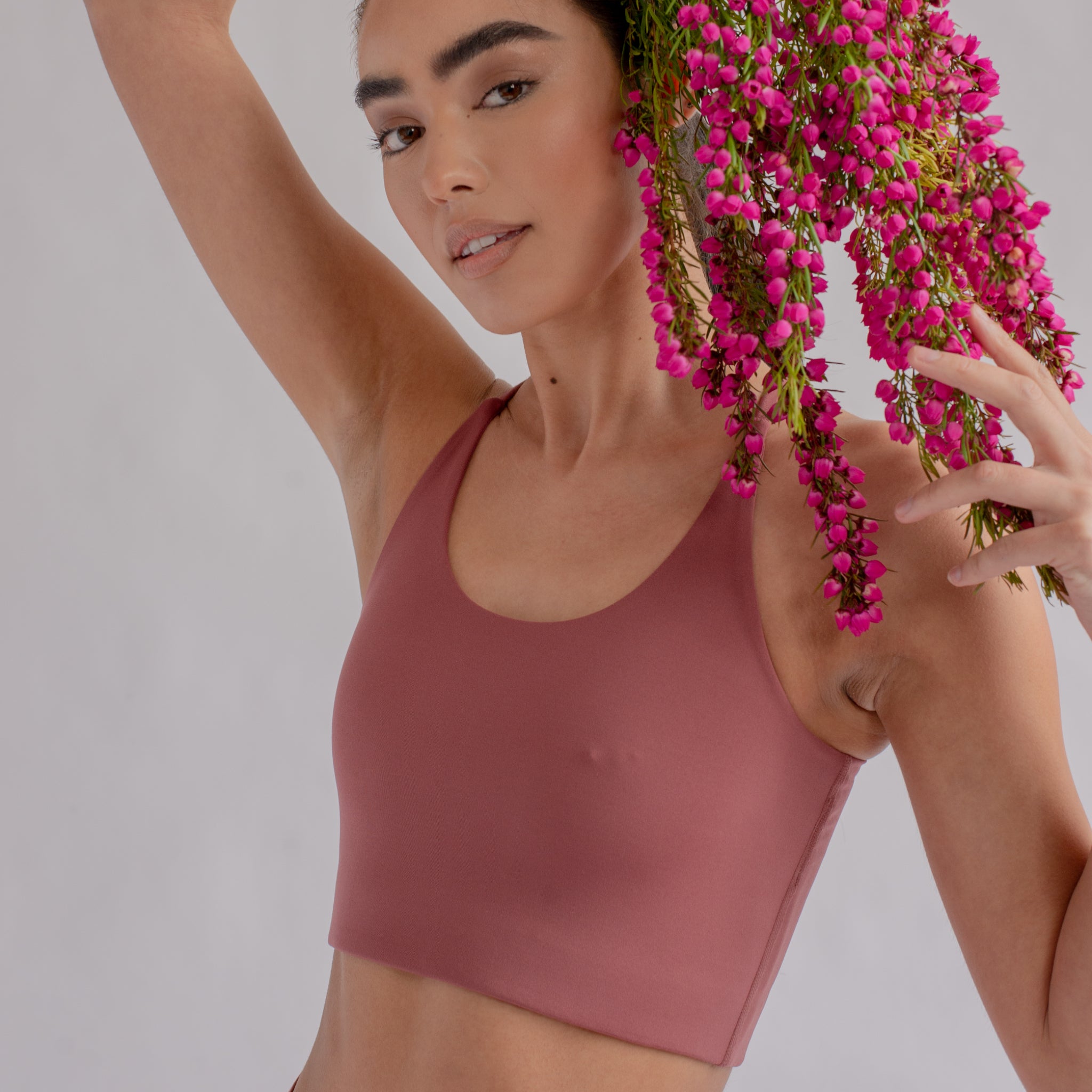 Frau in einem rosa Top hält einen Strauß aus pinken Blumen über ihrem Kopf, vor neutralem Hintergrund.