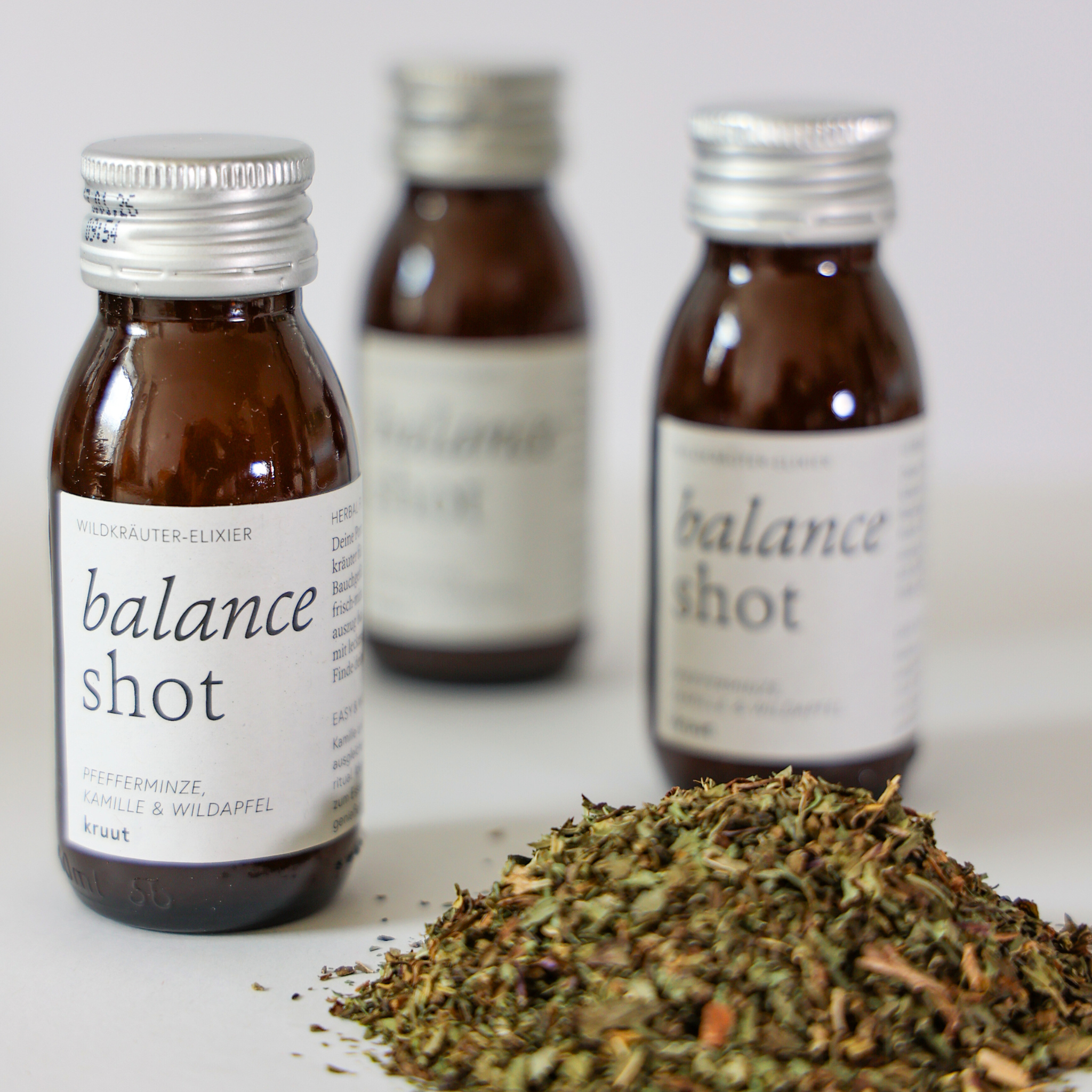 Drei braune Flaschen mit "balance shot"-Etikett und ein Haufen getrockneter Kräuter im Vordergrund.
