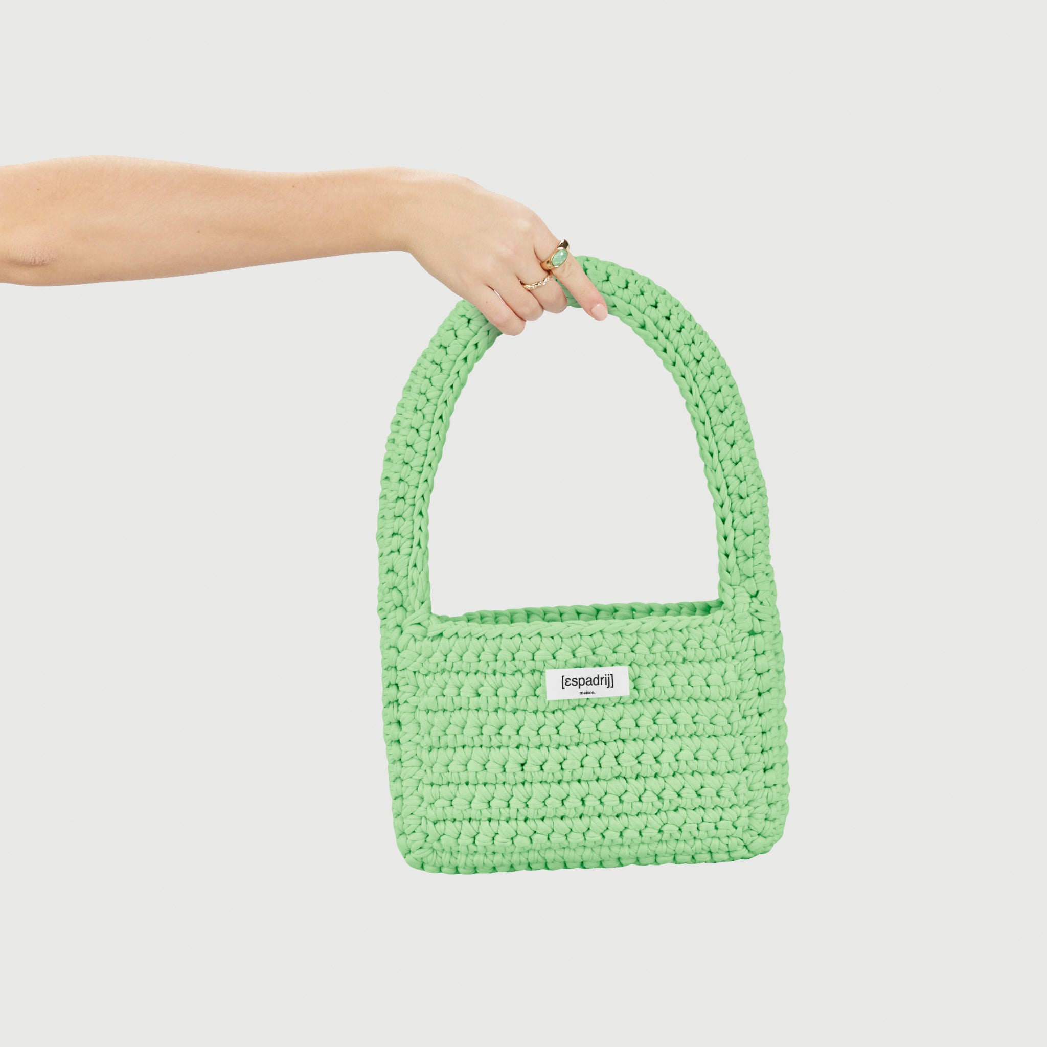 Ein Arm hält eine grüne, gestrickte Handtasche mit Label „espadrij“ vor weißem Hintergrund.