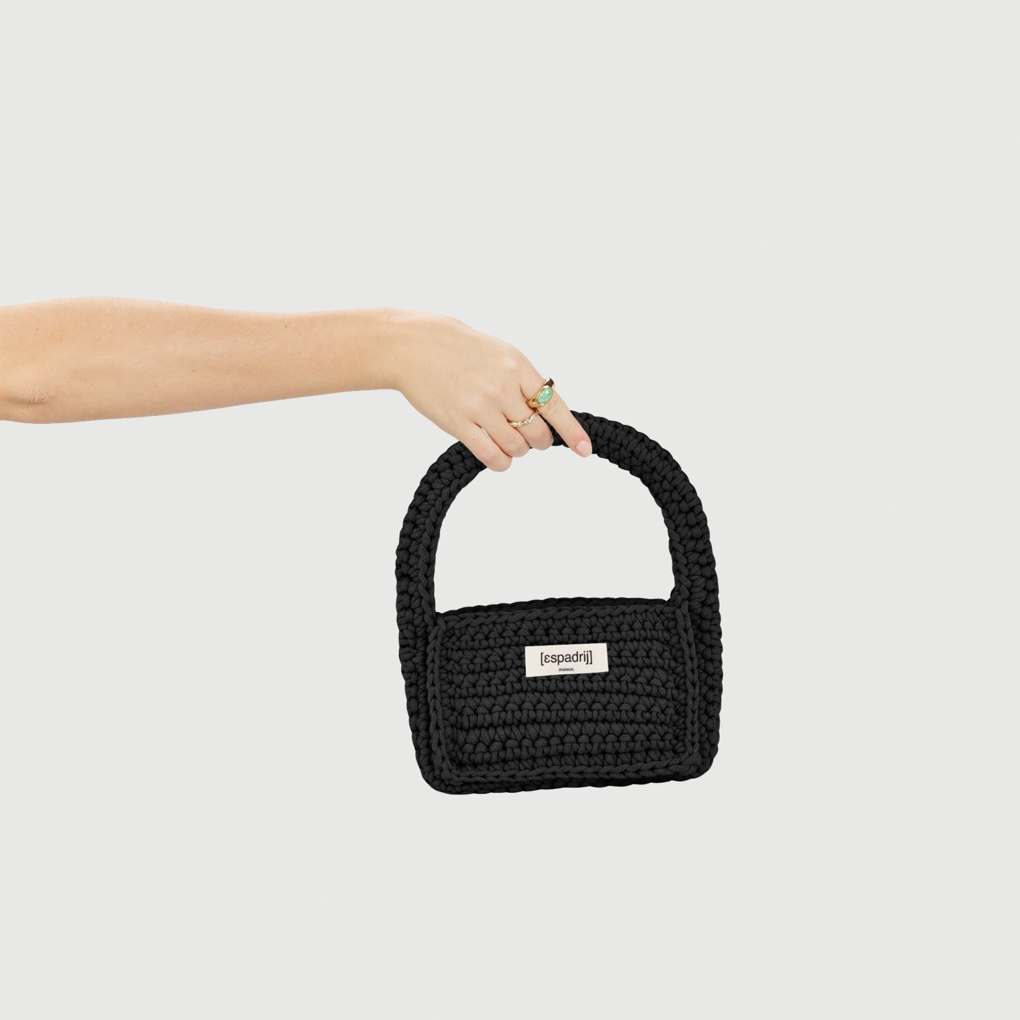 Eine Hand hält eine schwarze, gehäkelte Tasche mit einem Etikett, das "espadrij" zeigt, vor einem hellgrauen Hintergrund.