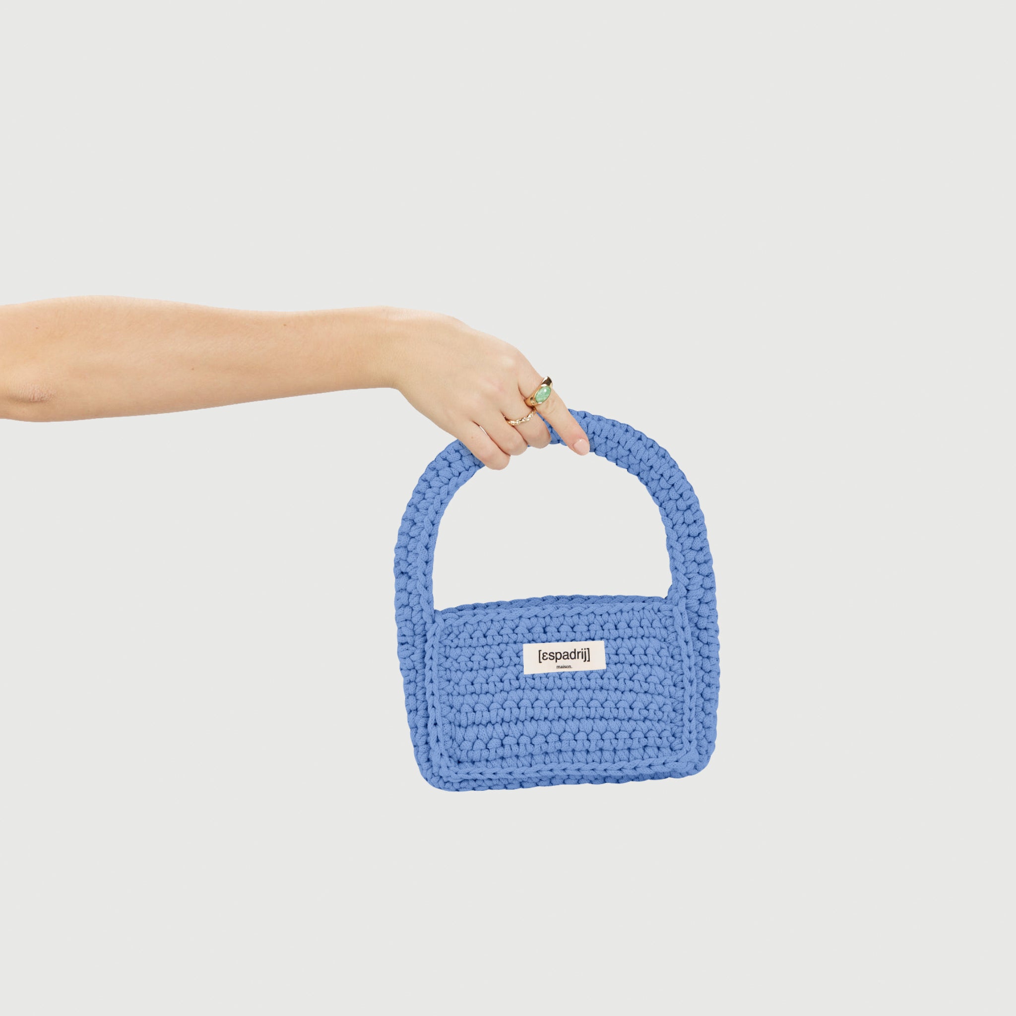 Eine Hand hält eine hellblaue, gestrickte Tasche mit einem weißen Label auf einem weißen Hintergrund.