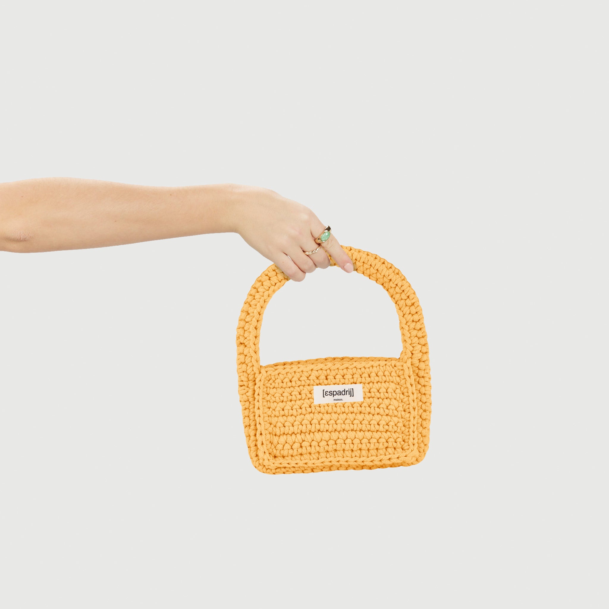 Eine Hand hält eine gelbe, gehäkelte Tasche mit einem kurzen Griff vor einem weißen Hintergrund.
