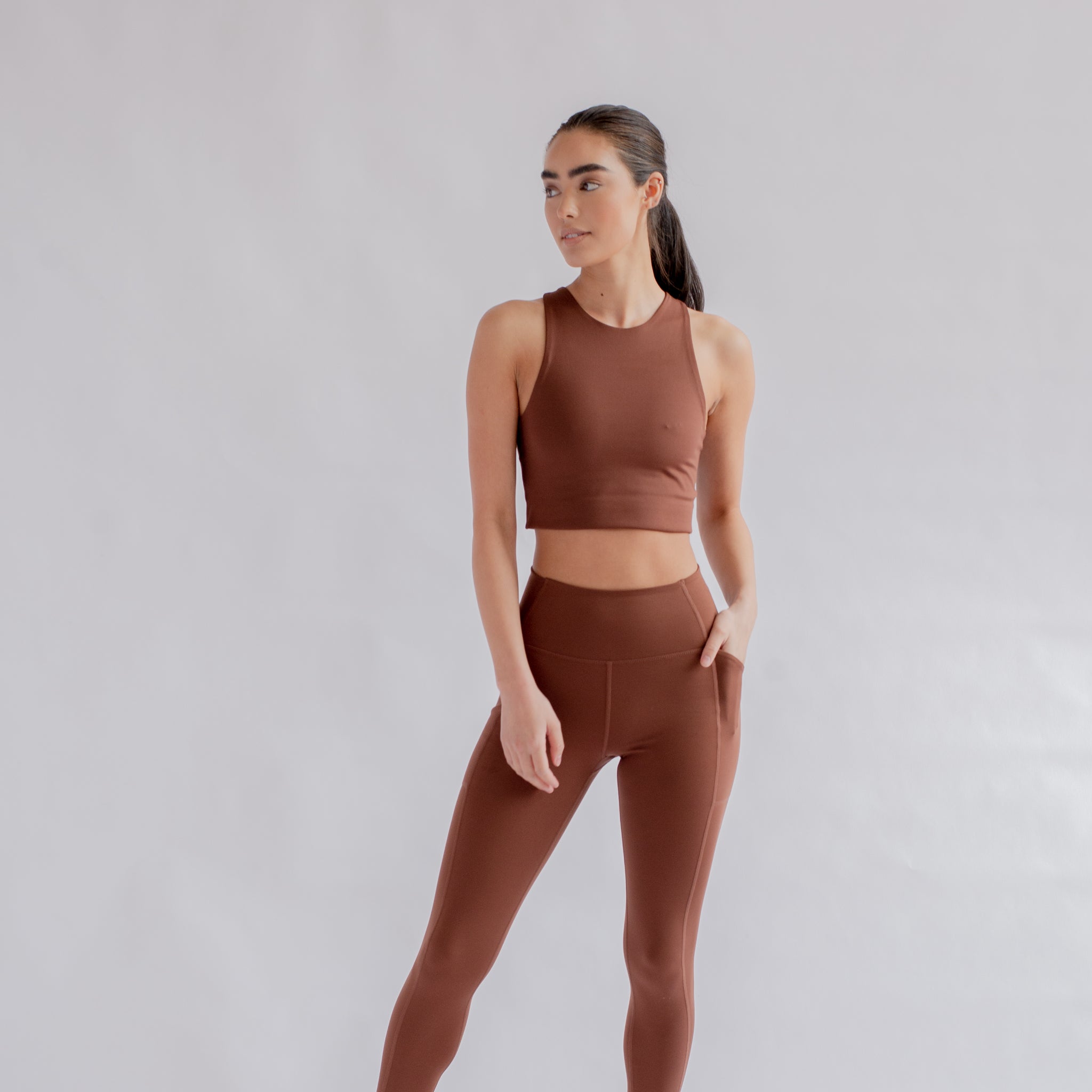 Person in brown athletic wear against neutral background, standing with one hand in pocket, looking to the side.