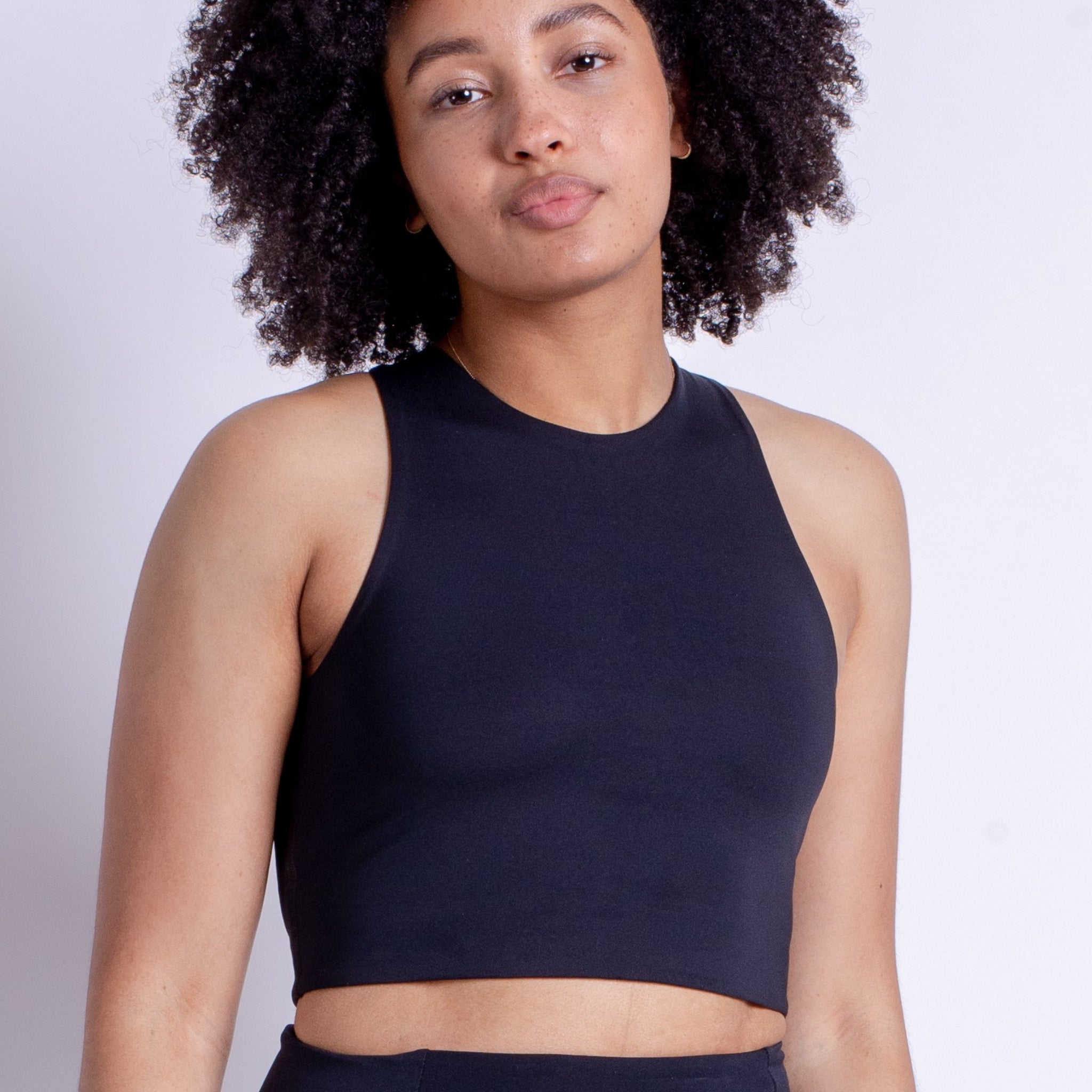 Eine Person mit lockigem Haar trägt ein schwarzes ärmelloses Oberteil vor einem weißen Hintergrund.