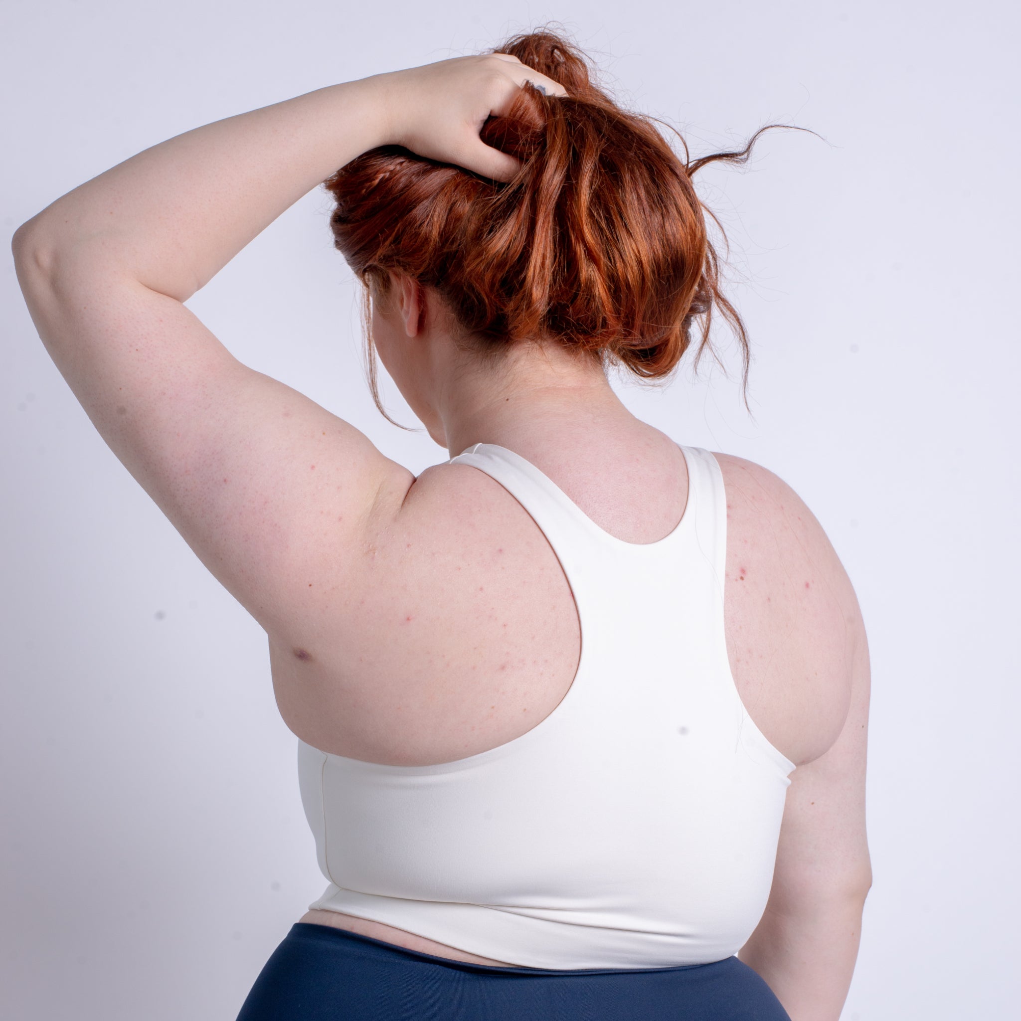 Eine Person mit roten Haaren in einem weißen Trägertop greift von hinten an ihren Kopf.