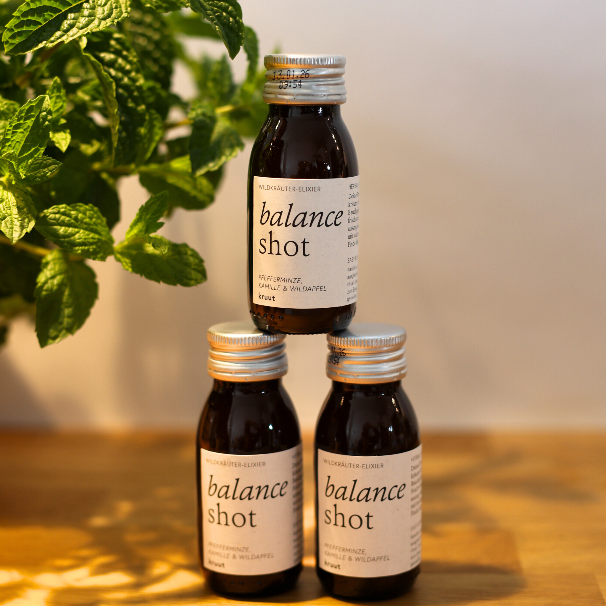 Drei Fläschchen mit der Aufschrift "Balance Shot" stehen gestapelt auf einem Tisch neben frischen Pfefferminzblättern.