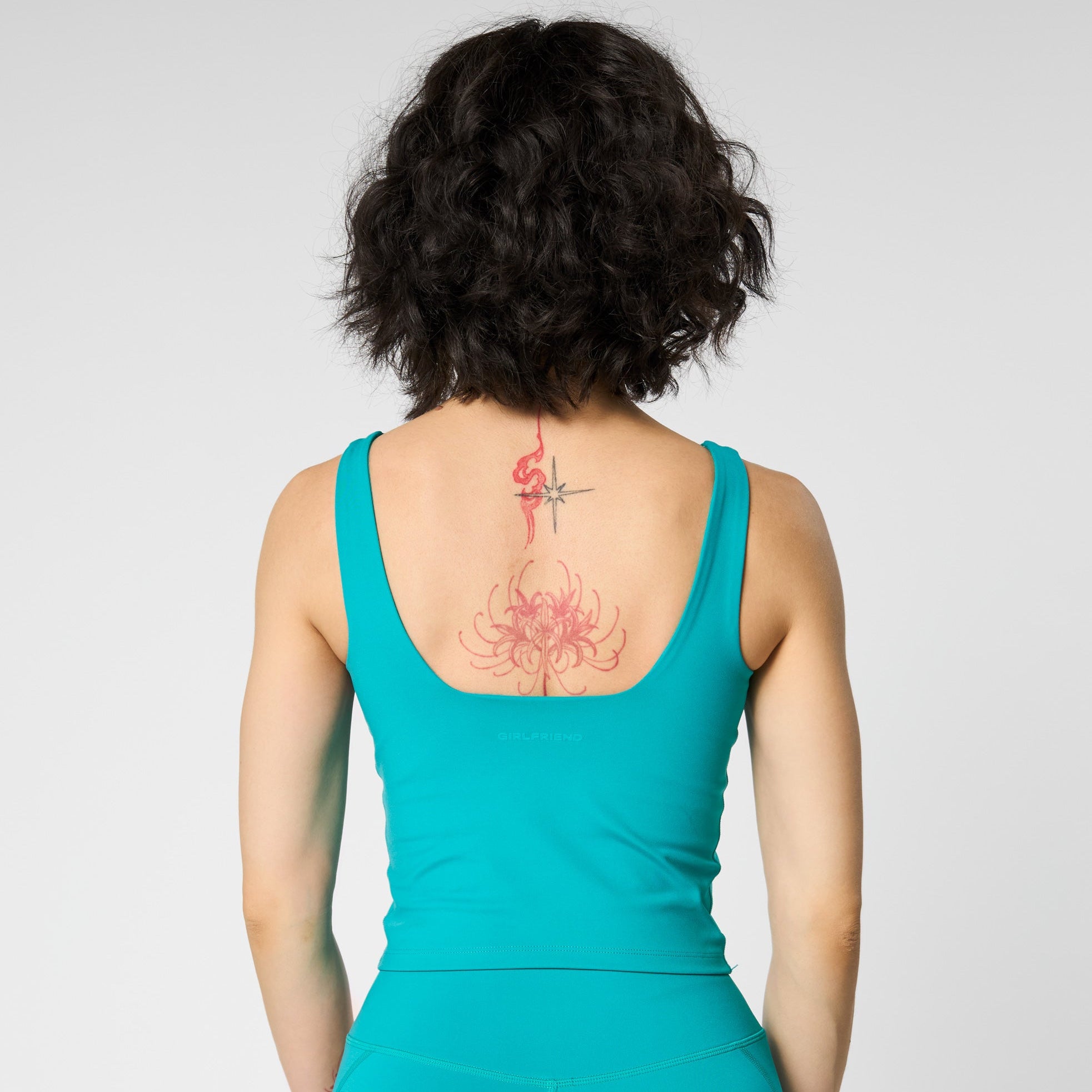 Eine Person mit kurzen, dunklen, gewellten Haaren steht mit dem Rücken zur Kamera und trägt ein blaugrünes, ärmelloses Oberteil und passende Shorts. Auf dem oberen Rücken ist eine rote Tätowierung zu sehen. Der Hintergrund ist einfarbig und hell.