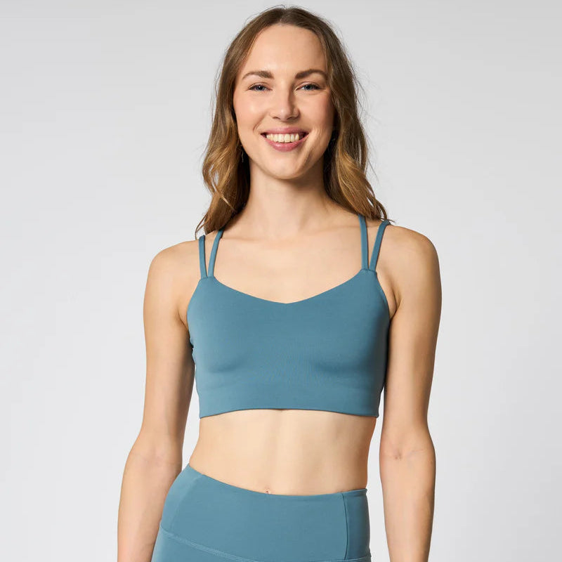 Eine Frau mit langen braunen Haaren lächelt, während sie einen passenden blauen Sport-BH und hoch taillierte Leggings trägt. Sie steht mit dem Gesicht nach vorne vor einem schlichten hellgrauen Hintergrund.