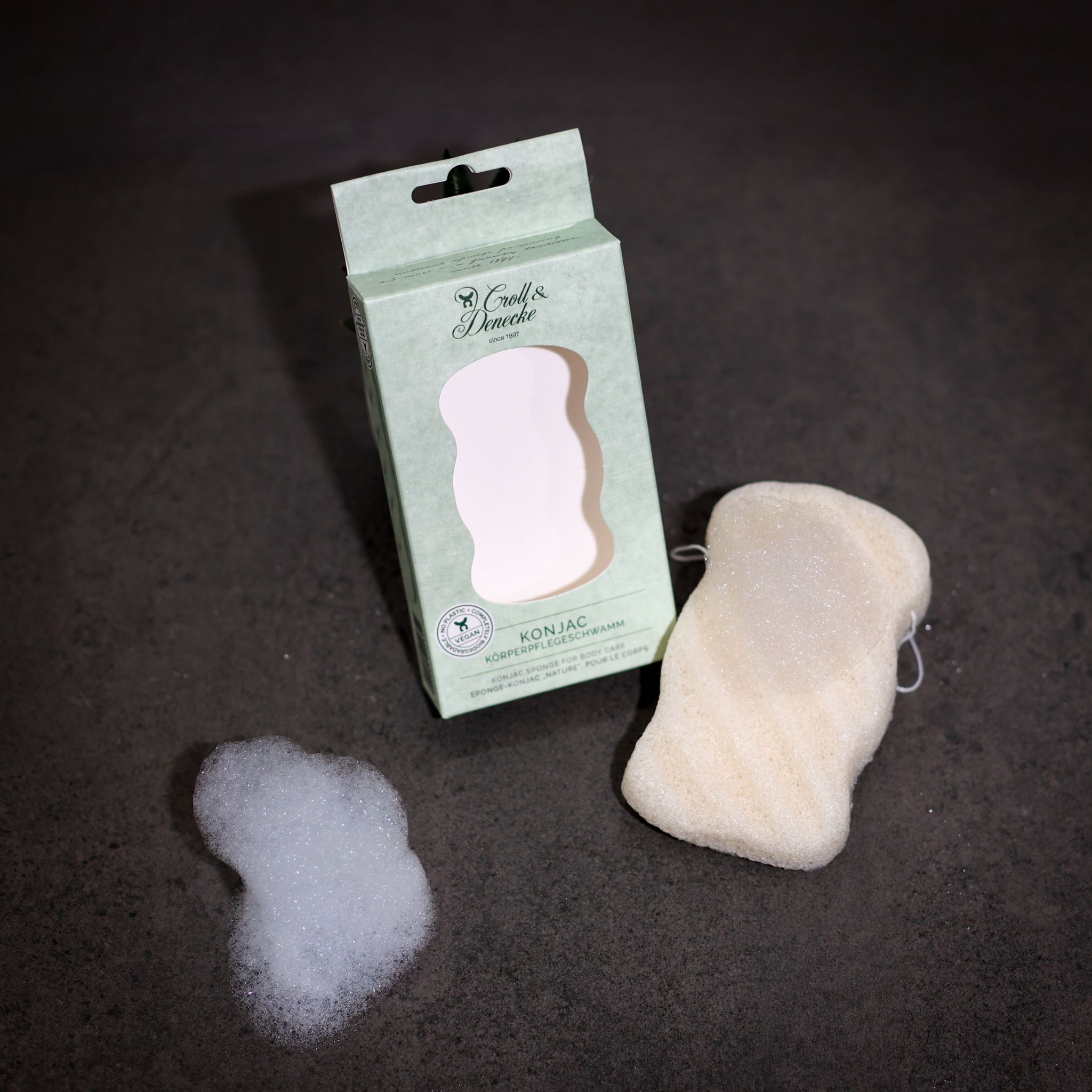 Ein Konjac-Körperschwamm mit Verpackung und etwas Schaum auf einem dunklen Untergrund.