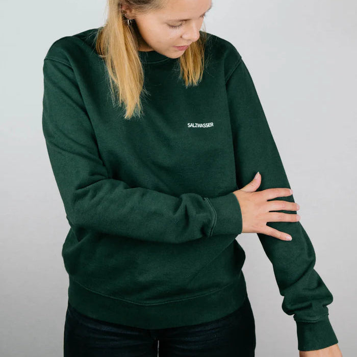 Eine Frau, die ein dunkelgrünes Sweatshirt mit dem Aufdruck SALZWASSER auf der Brust trägt, blickt nach unten und berührt ihren Ärmel vor einem schlichten hellen Hintergrund.