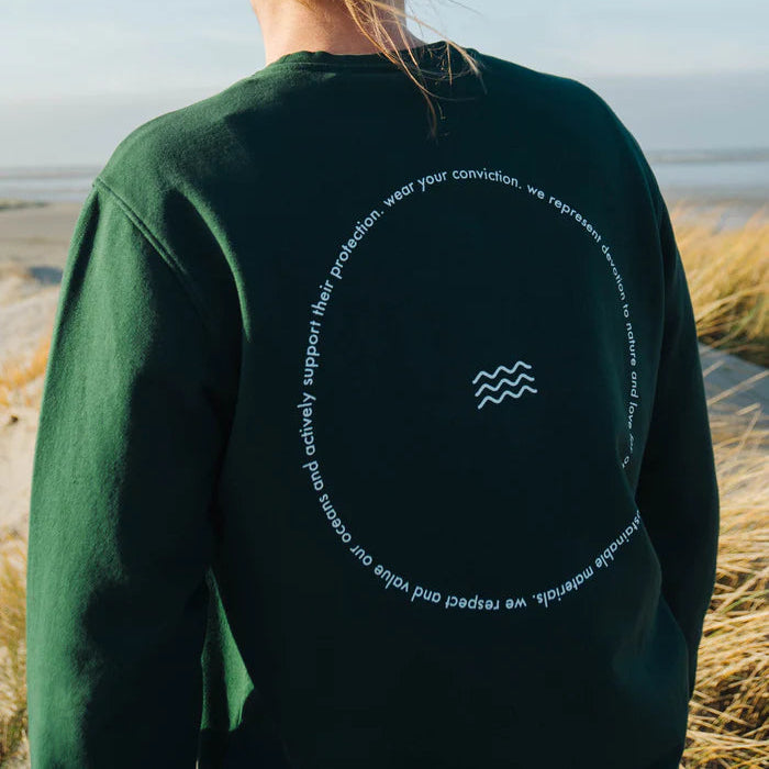 Eine Person, die ein dunkelgrünes Sweatshirt trägt, steht im Freien in der Nähe von hohem Gras. Auf der Rückseite des Sweatshirts ist ein kreisförmiger Text abgebildet, der ein kleines Wellensymbol in der Mitte umgibt. Die Szene scheint an einem Strand oder Küstengebiet zu spielen.