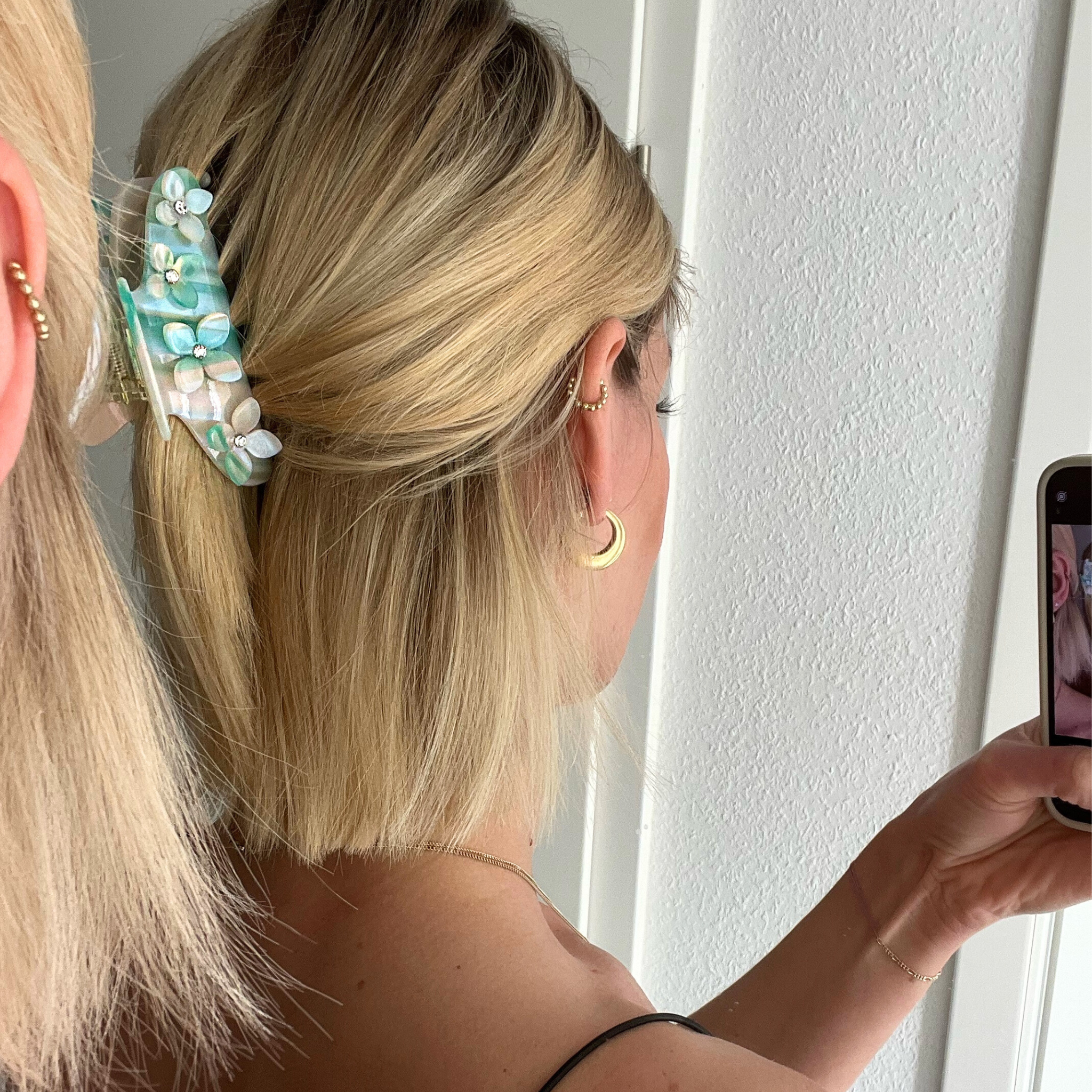 Frau mit blonden Haaren und blauer Haarspange macht ein Spiegel-Selfie, trägt goldene Ohrringe und Ohrschmuck.