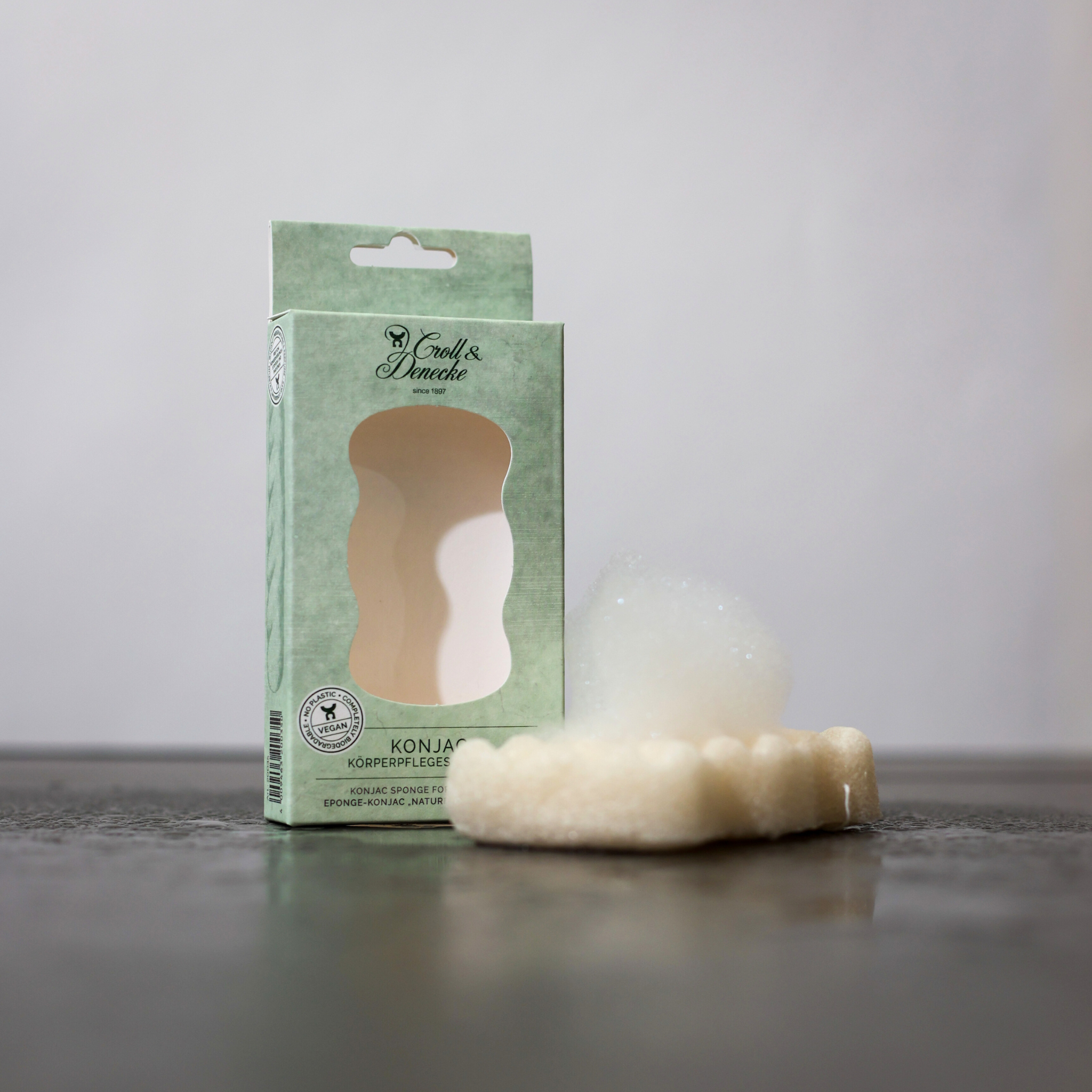 Eine Konjak-Körperpflege-Schwammverpackung steht neben einem schaumigen Schwamm auf einer dunklen Oberfläche.